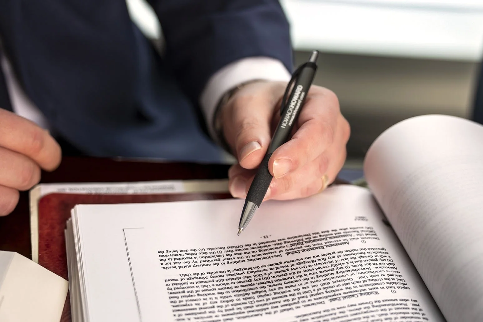 Close-up of a person's hand holding a black pen with the word 'ON' in white, writing or signing on a thick book or legal document with dense text, on a dark brown desk.