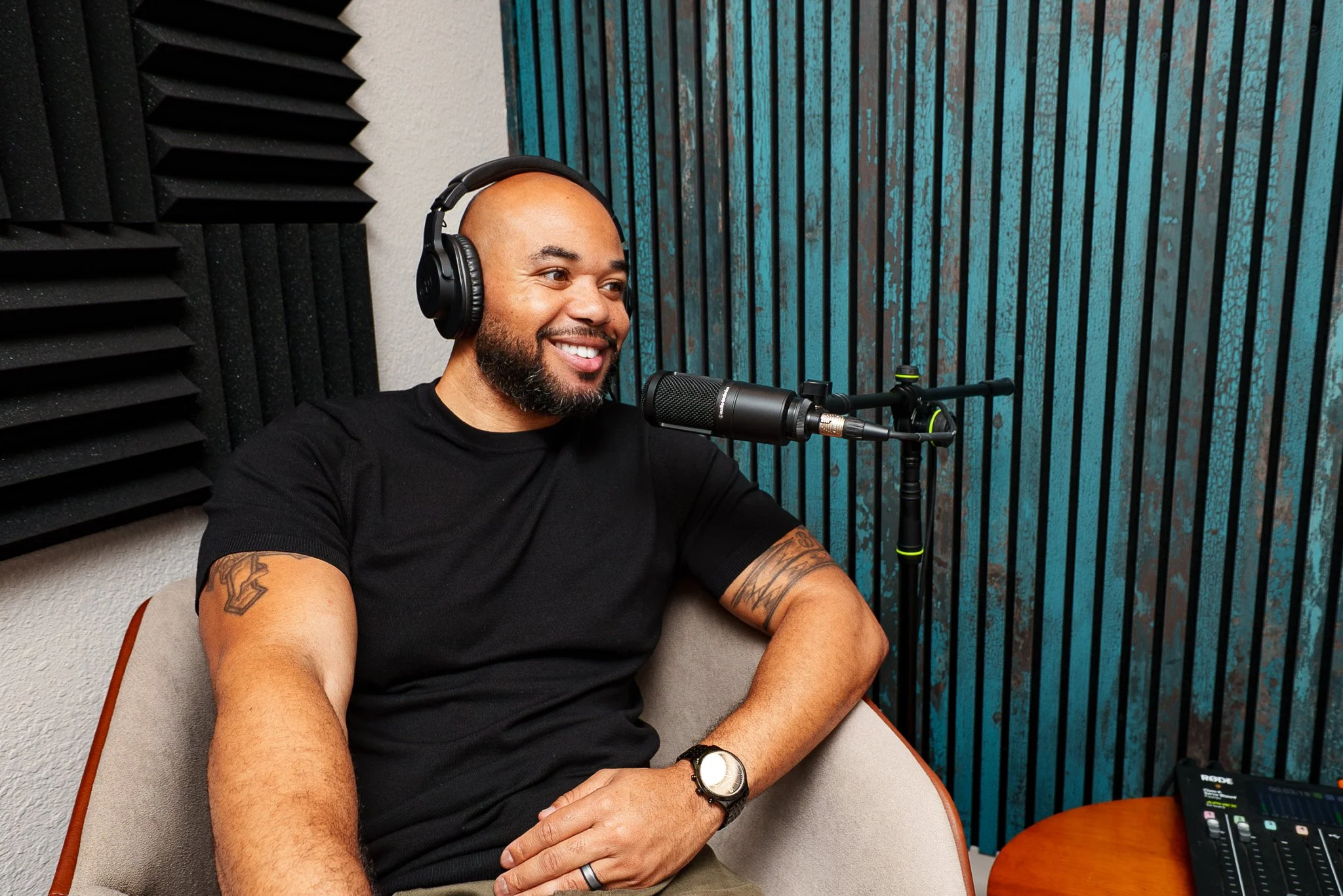 A man with a beard and tattoos, wearing a black t-shirt and headphones, smiles while speaking into a microphone in a podcast studio.