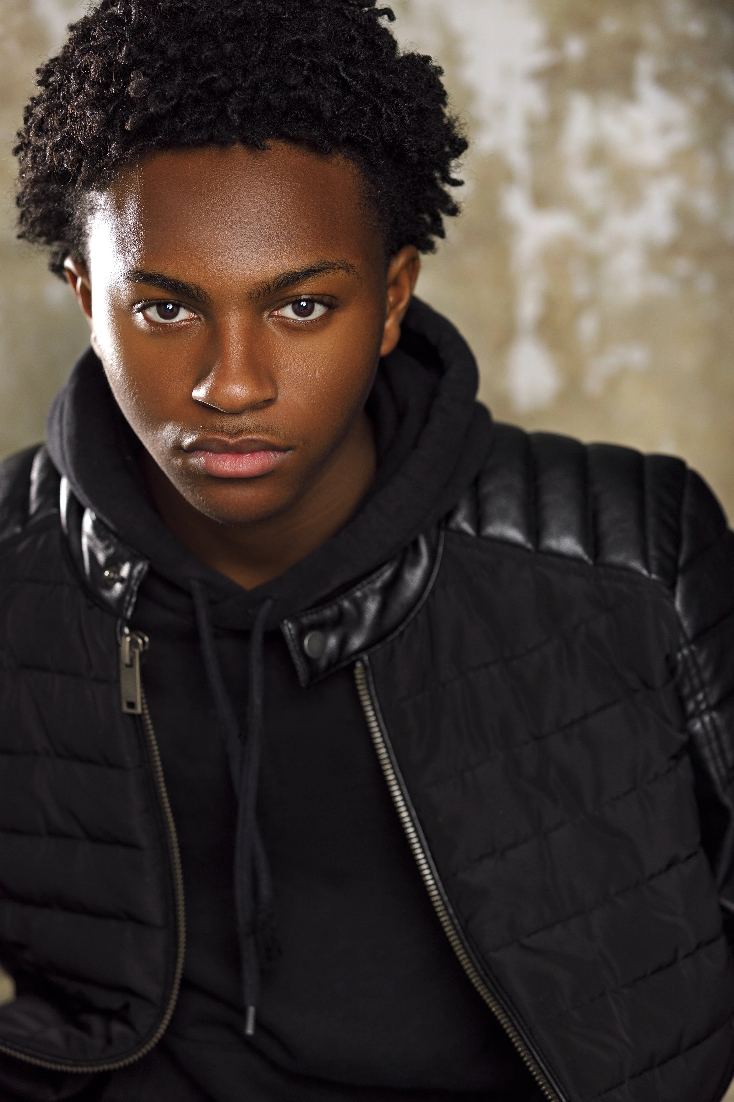 A young African American woman with short curly hair and a serious expression, wearing a black quilted jacket and hoodie, looking directly at the camera.