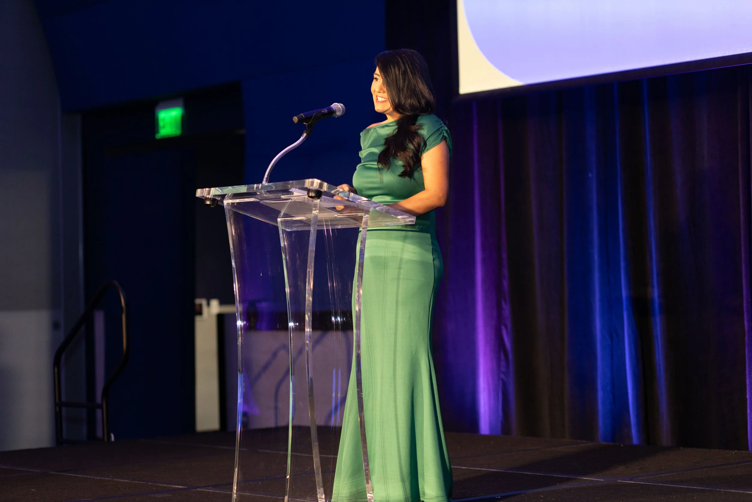 A woman in a green dress standing at a clear podium, speaking into a microphone on stage with purple curtains in the background.