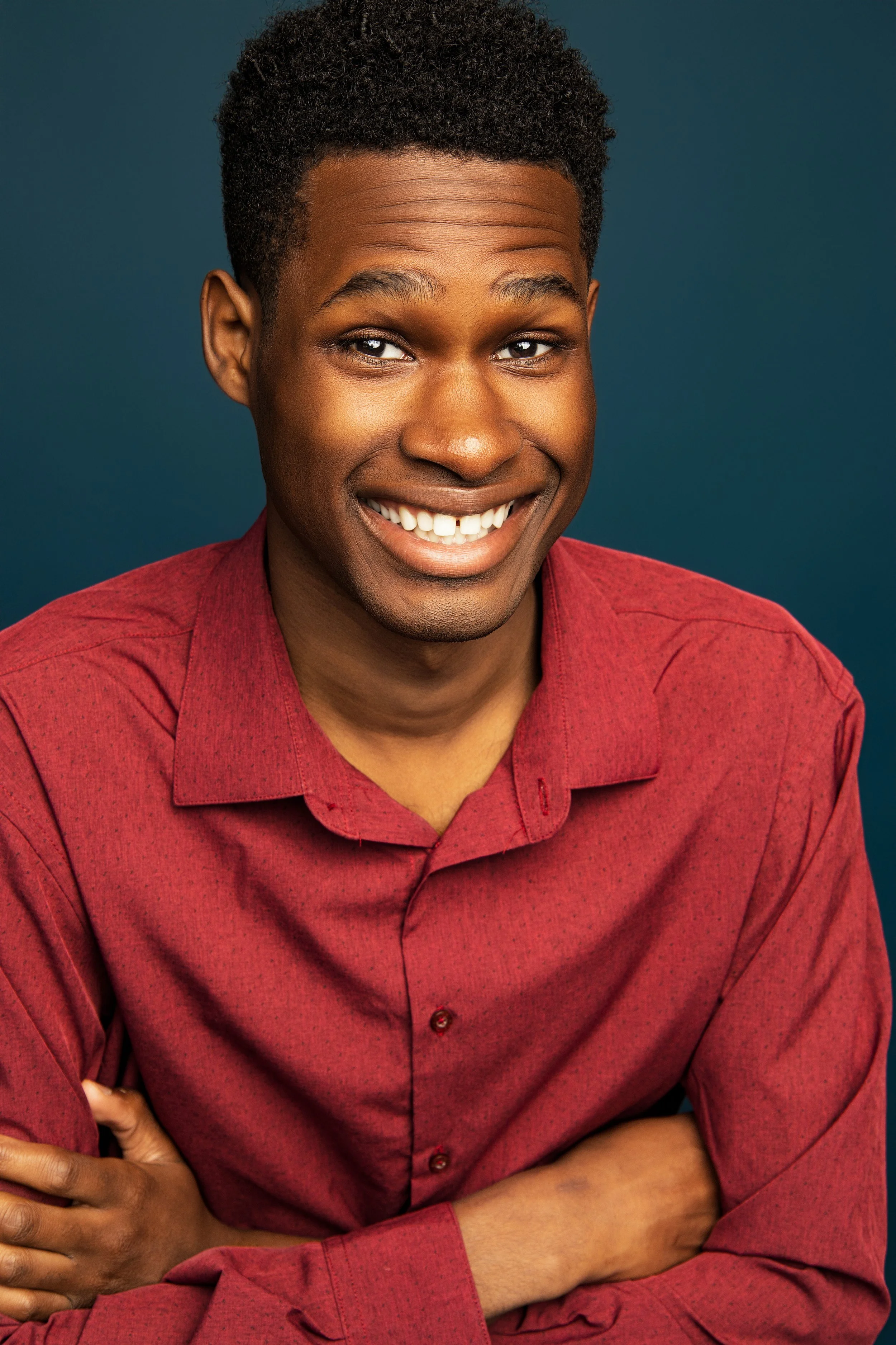 A young man smiling, wearing a red shirt, with arms crossed, against a dark blue background.