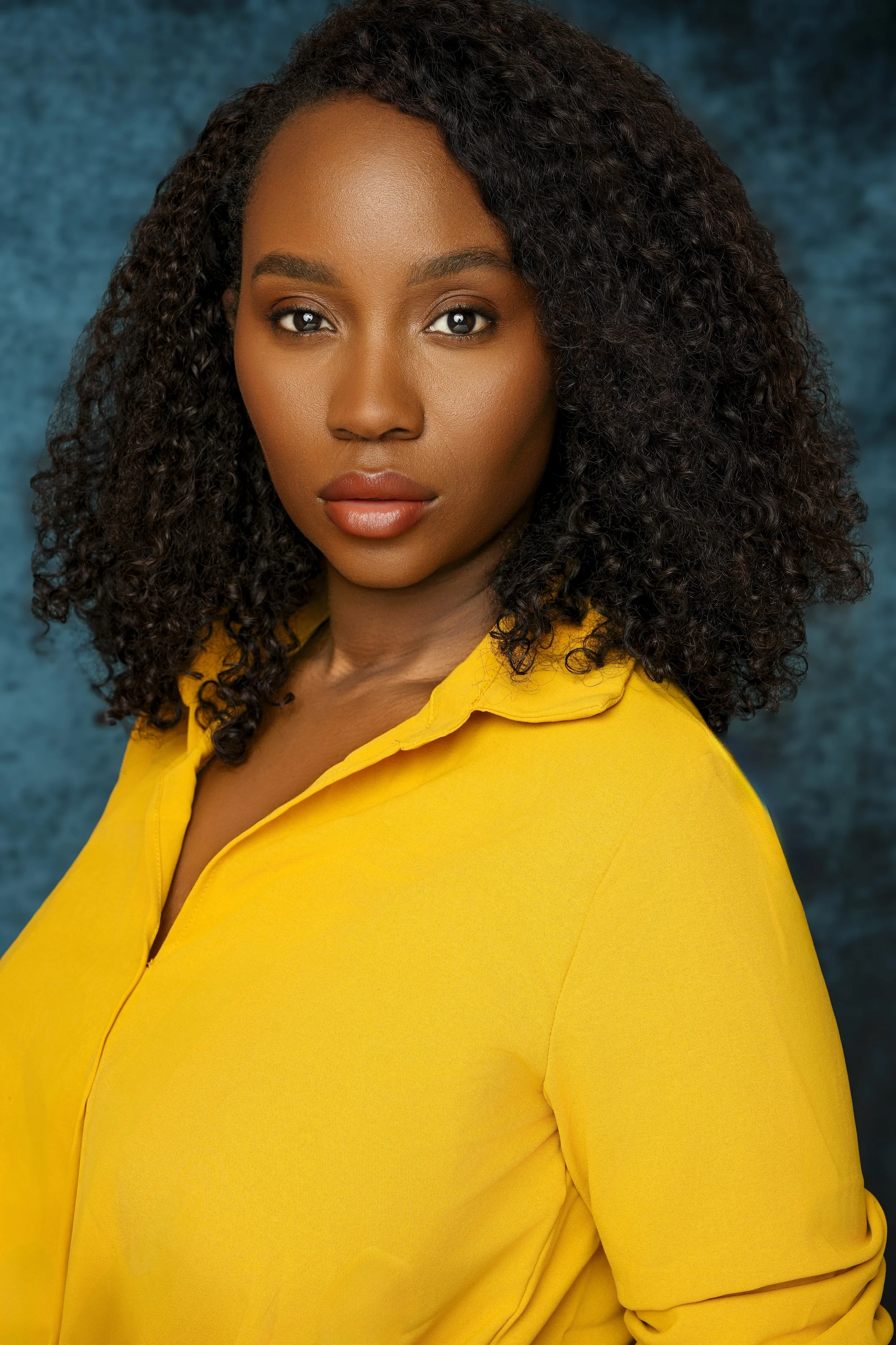 Portrait of a woman with curly hair wearing a yellow top against a textured blue background.