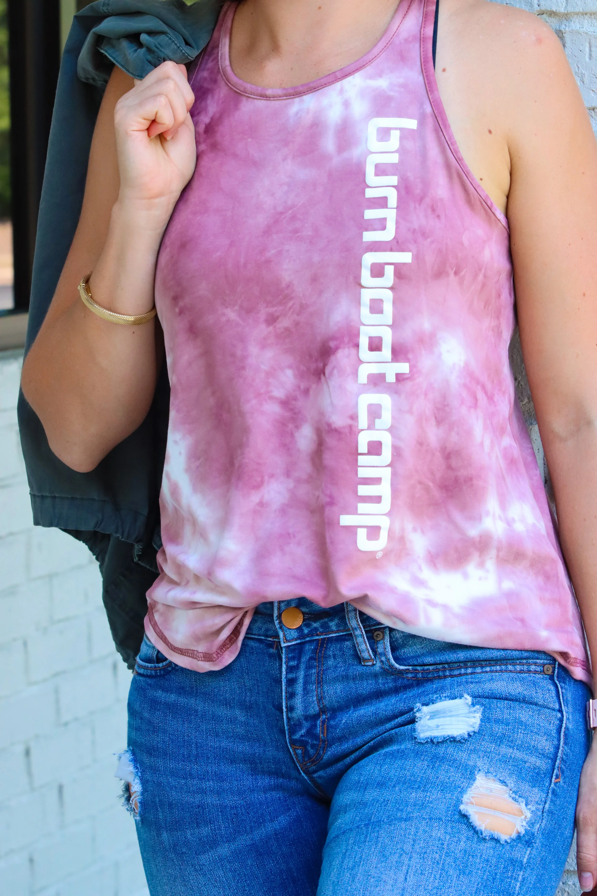 Person wearing a pink tie-dye tank top with white text reading 'run barefoot' and ripped blue jeans, holding a jacket over shoulder.