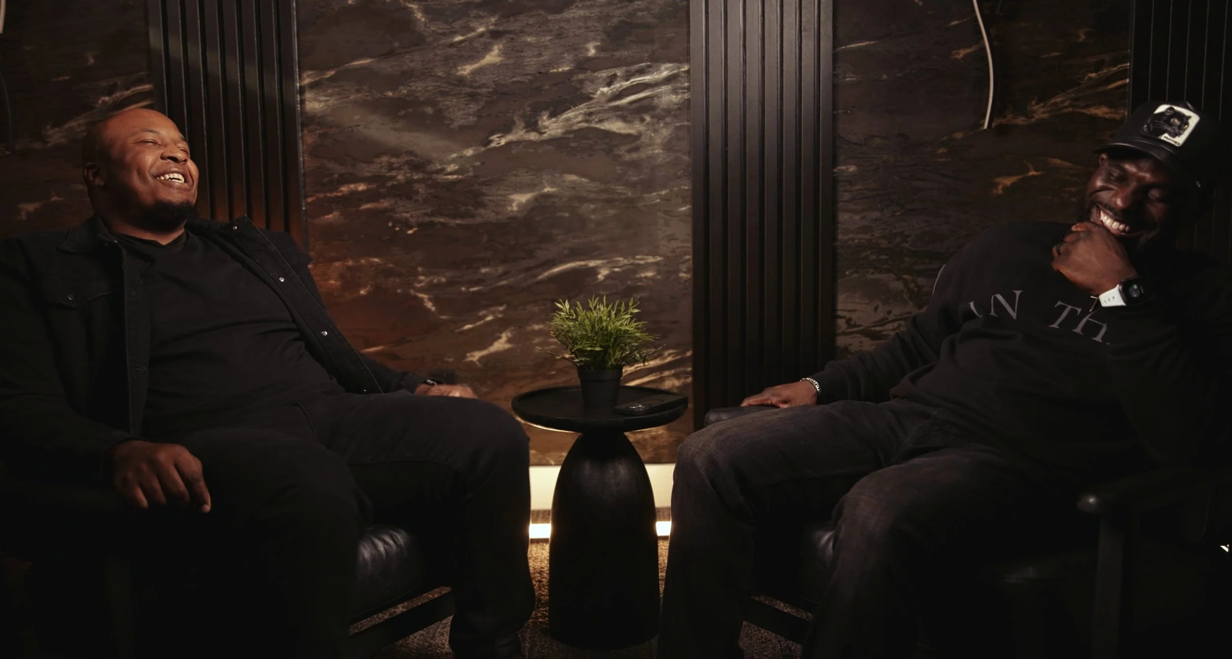 Two men sitting and laughing in a dimly lit room, with a marble-patterned wall behind them. There is a small black table with a green plant between them.