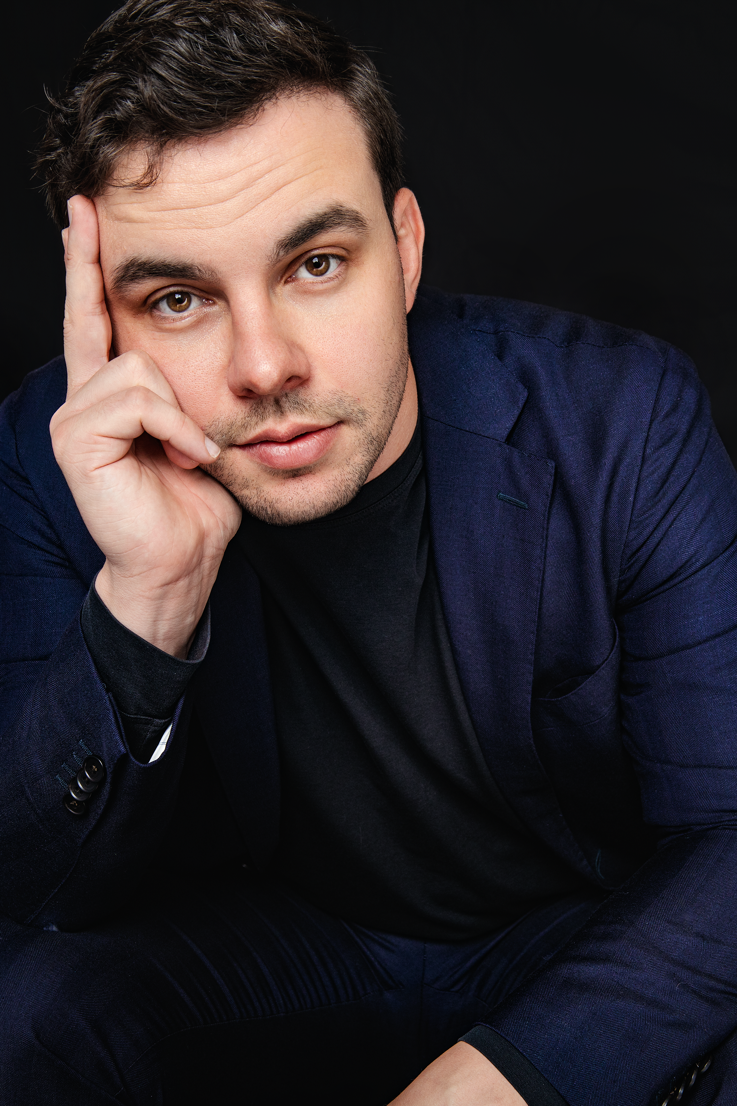 A man with brown hair and brown eyes posing against a black background, wearing a dark blue blazer and black shirt, looking into the camera with his hand resting on his face.