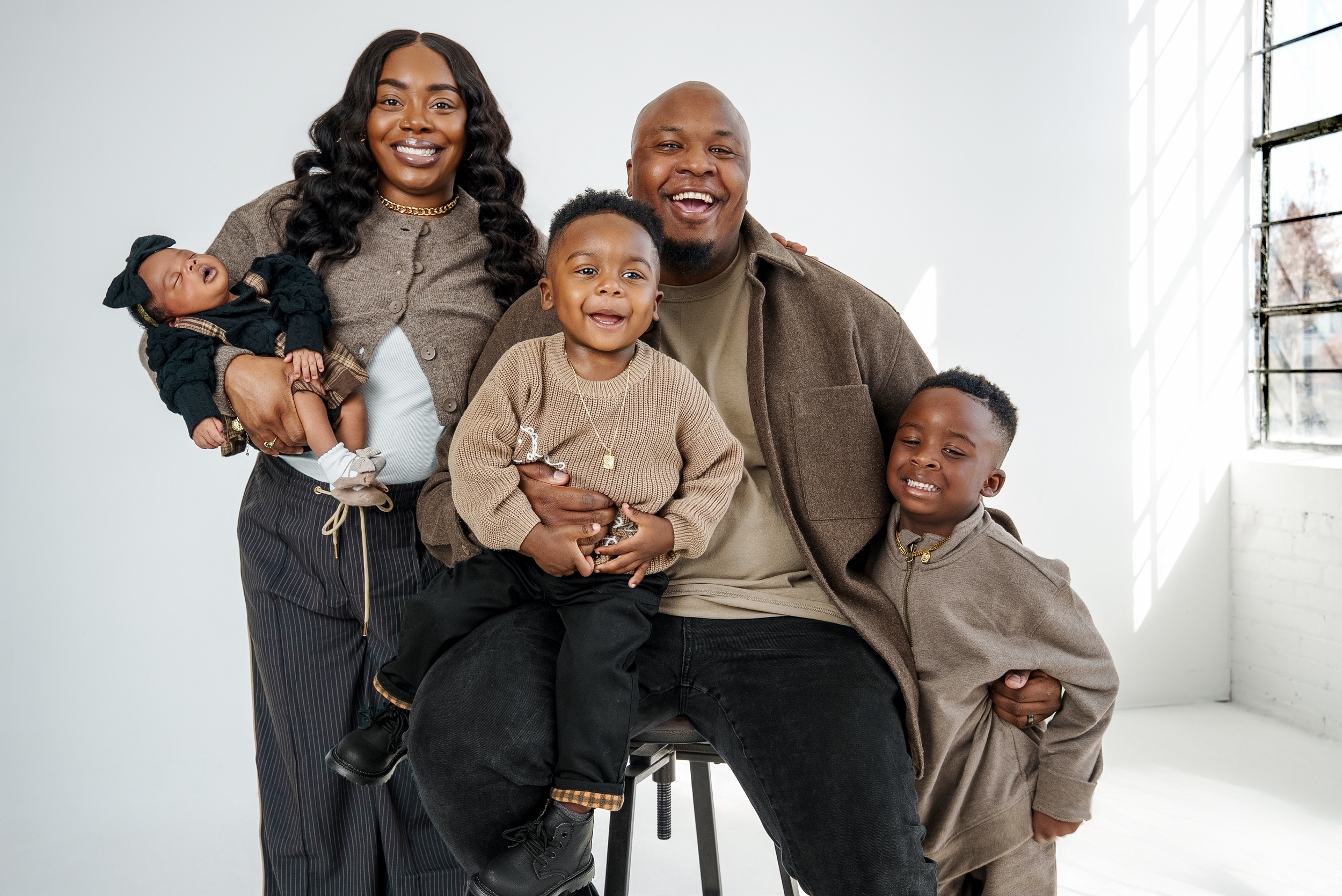 A happy family of six posing together indoors near a window with white walls. The woman on the far left holds a sleeping baby girl. The man in the center, seated, holds a young boy on his lap and has another boy standing next to him. The woman has lo