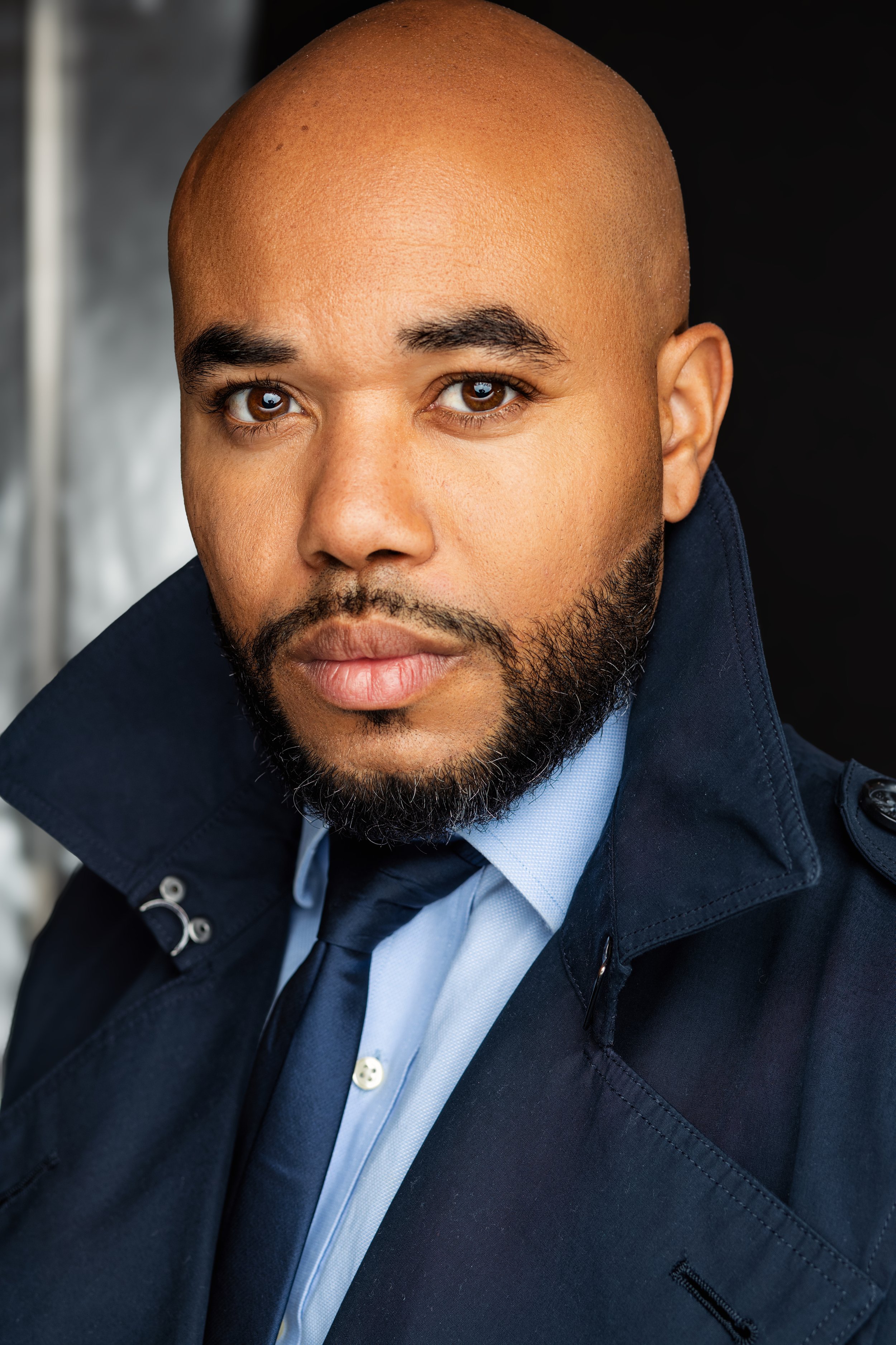 A close-up portrait of a man with a bald head, brown eyes, and a beard, wearing a dark navy coat and a light blue shirt with a dark tie, looking directly at the camera.