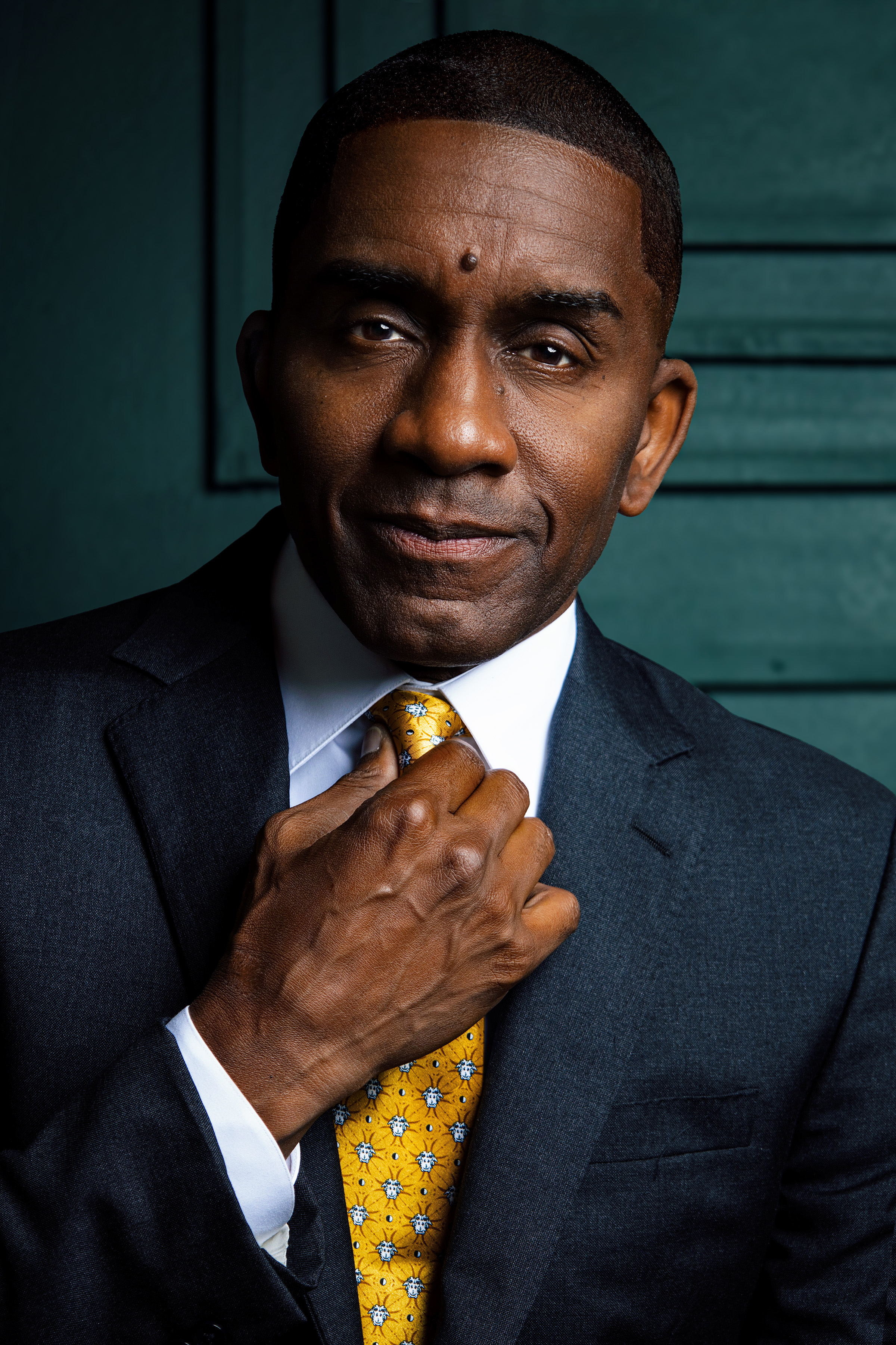A middle-aged man in a dark suit and yellow patterned tie adjusting his tie, standing in front of a teal-colored wall.
