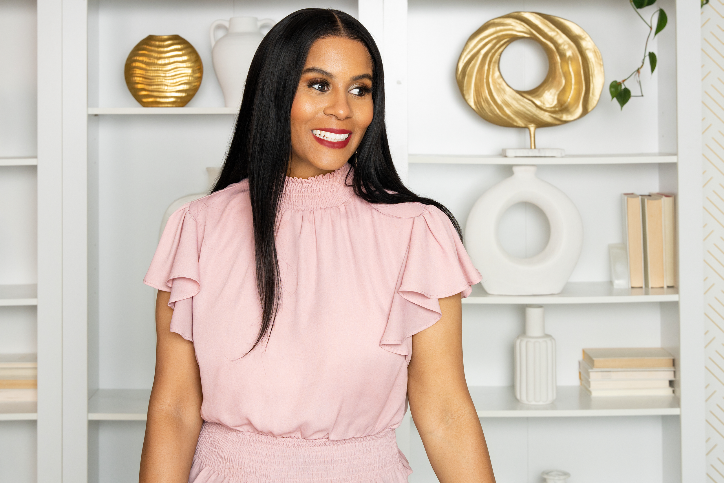 A woman with long dark hair wearing a light pink blouse with ruffled sleeves, smiling and looking to her left, in front of a white bookshelf with decorative vases and books.