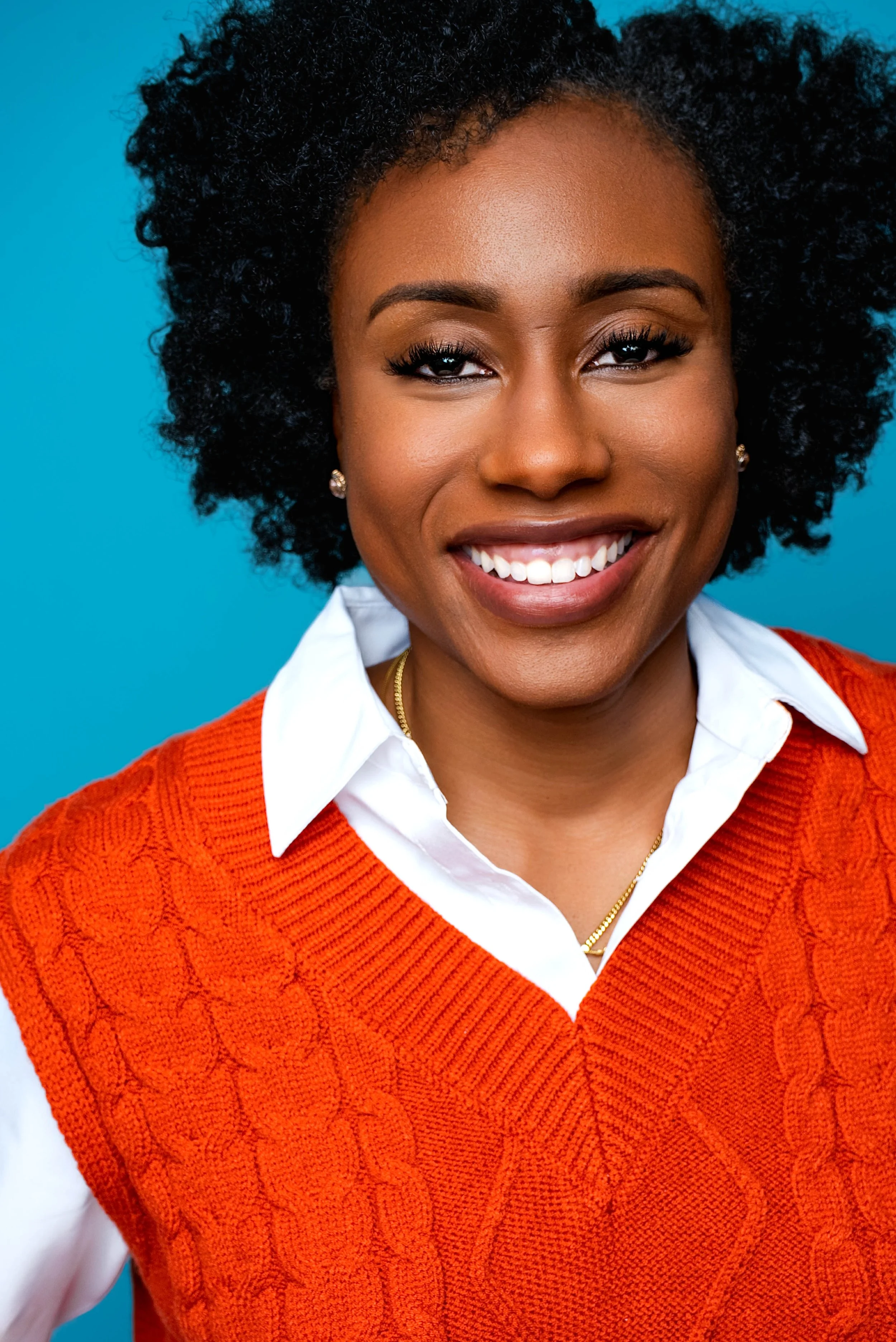 A smiling Black woman with curly hair, wearing a white collared shirt under an orange sweater, against a blue background.