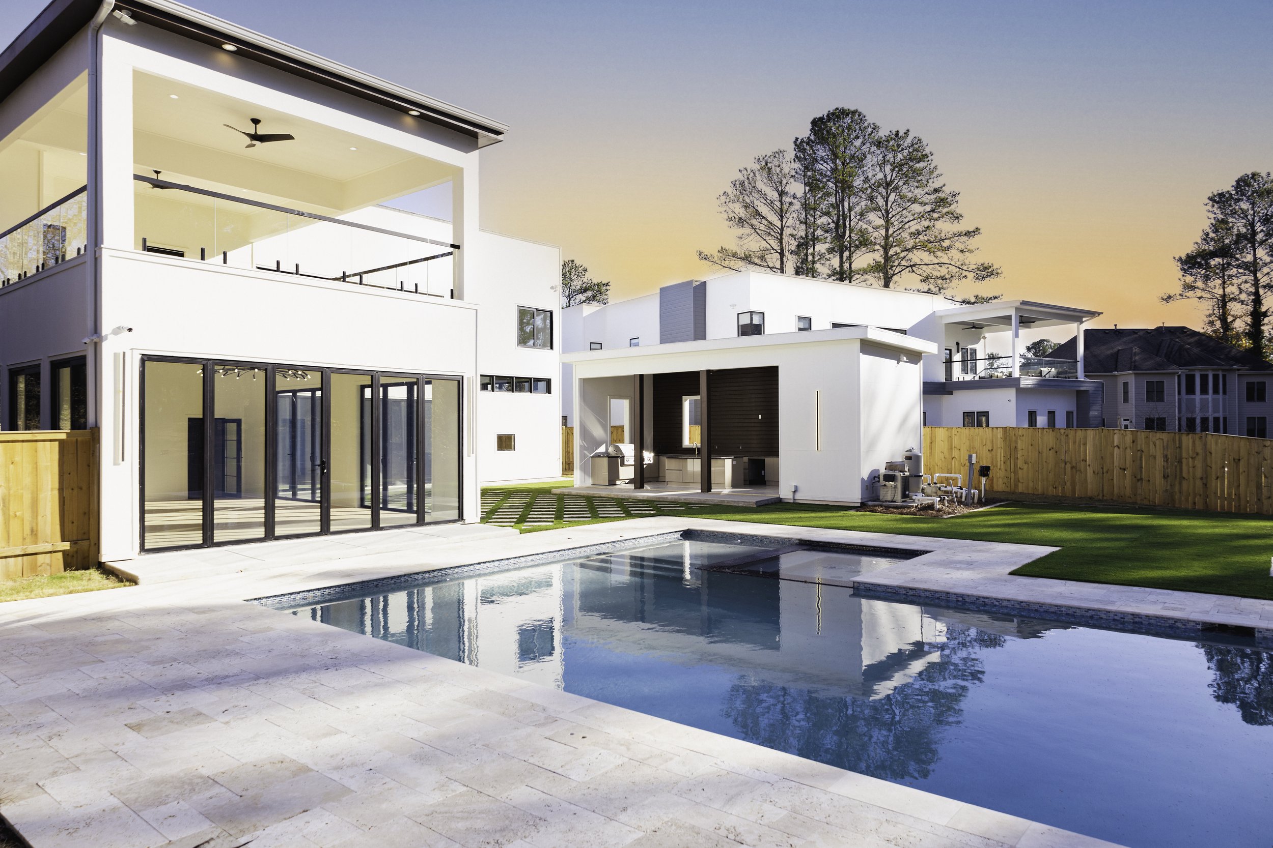 Modern house with white exterior, large glass doors, swimming pool, and a fenced backyard at sunset.