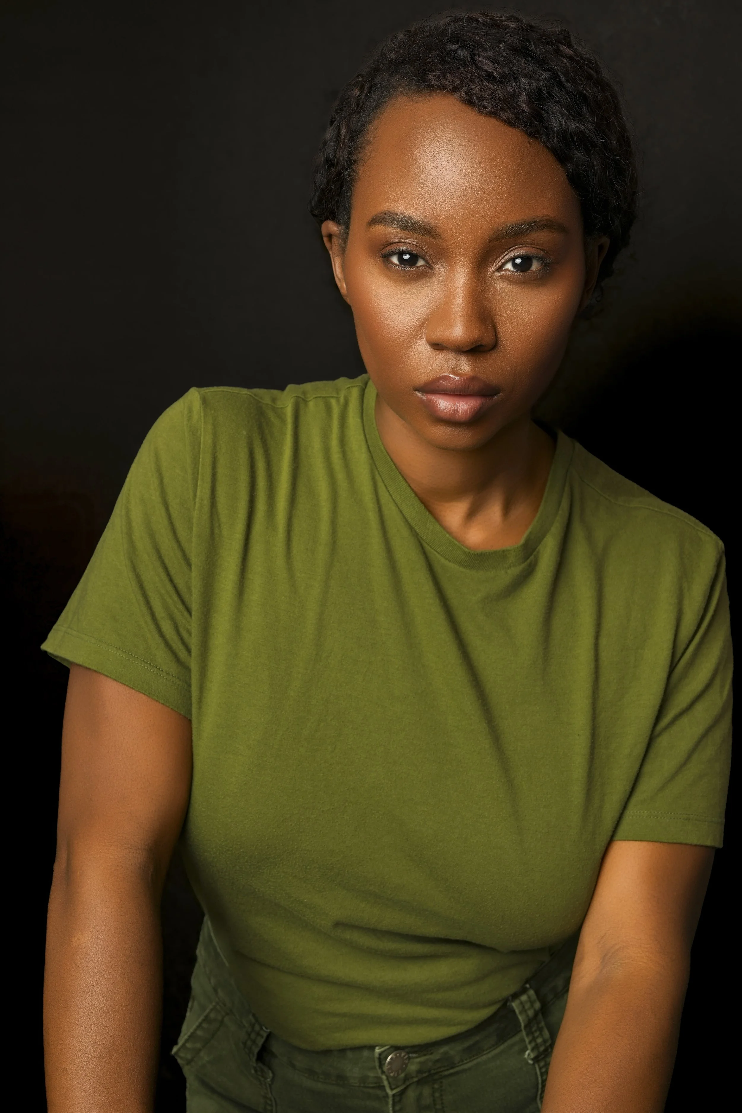 A young woman with short curly hair, wearing an olive green T-shirt, looking directly at the camera against a black background.