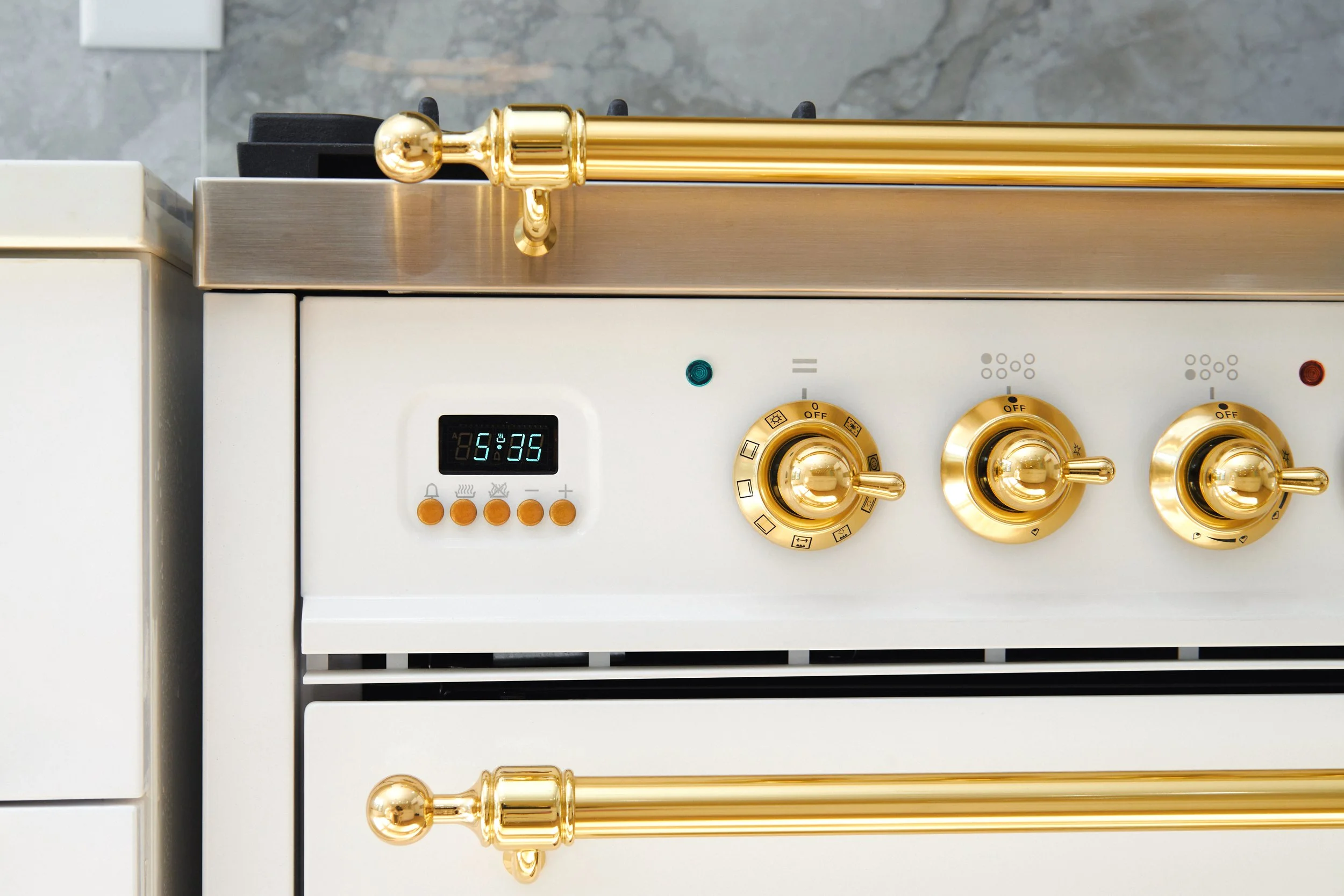 Close-up of a white stove with golden knobs and a digital clock showing 5:35, with a marble countertop and gray wall in the background.