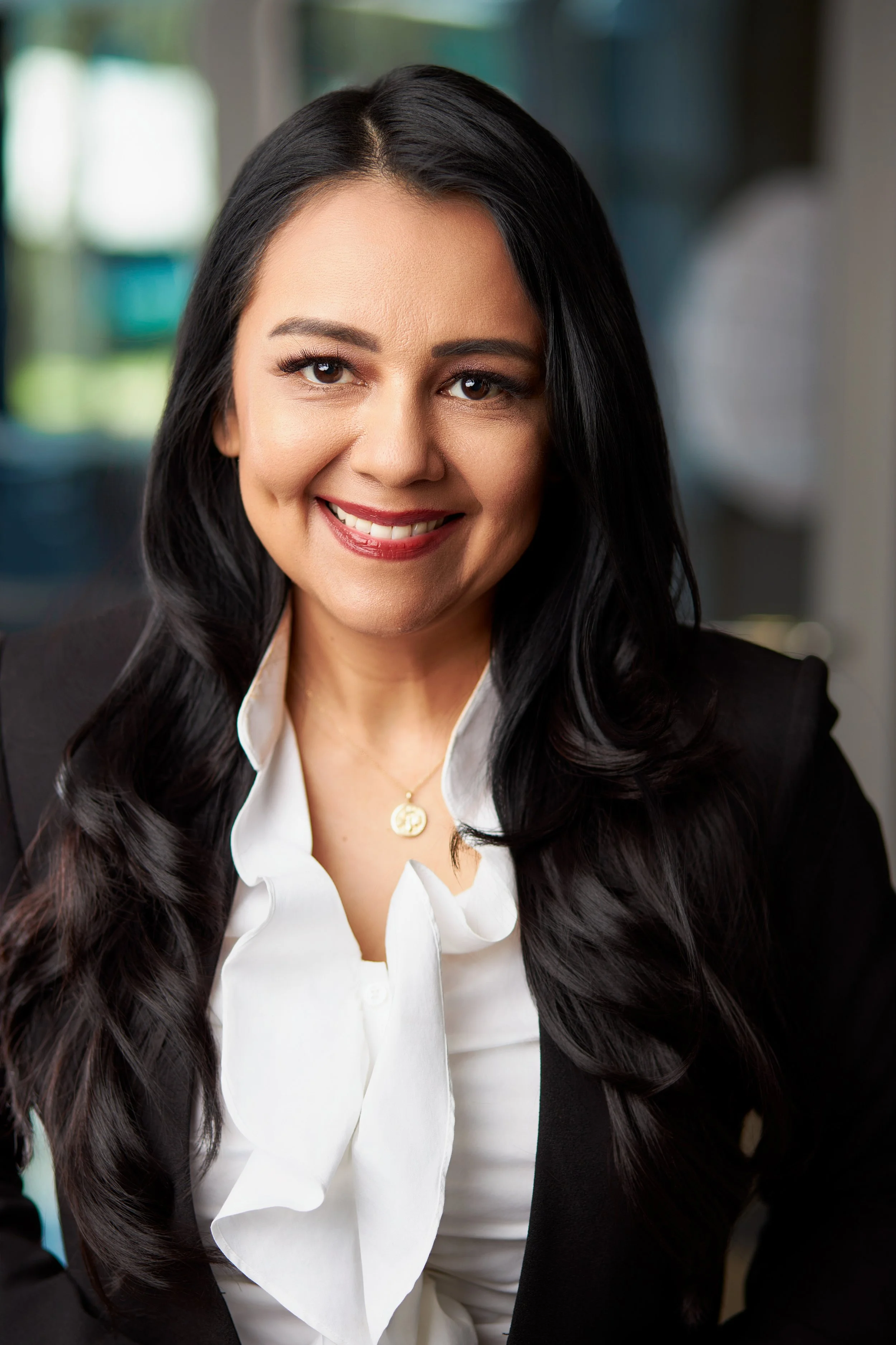 A smiling woman with long black hair wearing a black blazer and white blouse, sitting indoors.