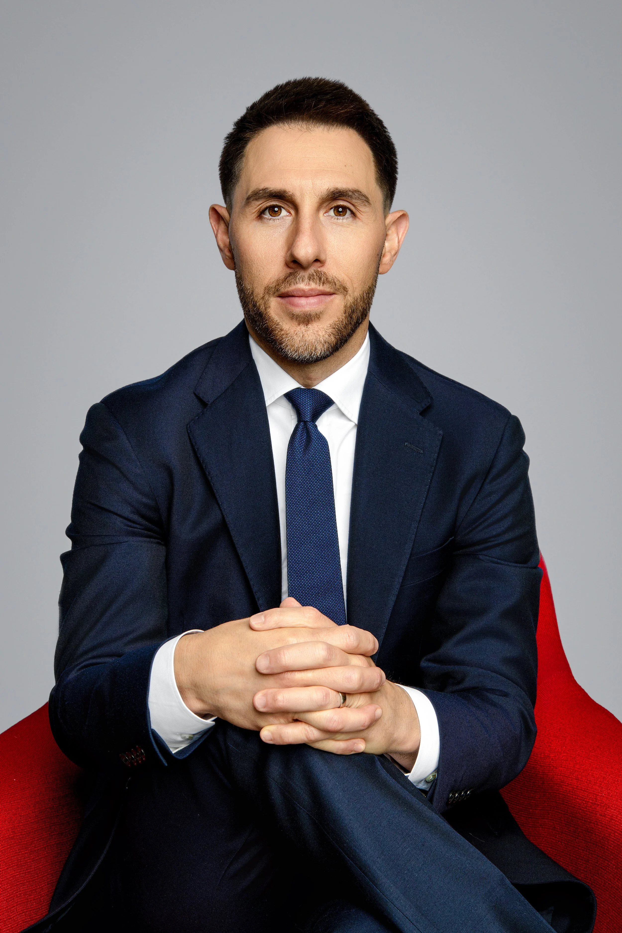 A man in a navy blue suit with a white shirt and matching tie, sitting on a red chair against a gray background, with hands clasped on his lap and a neutral expression.