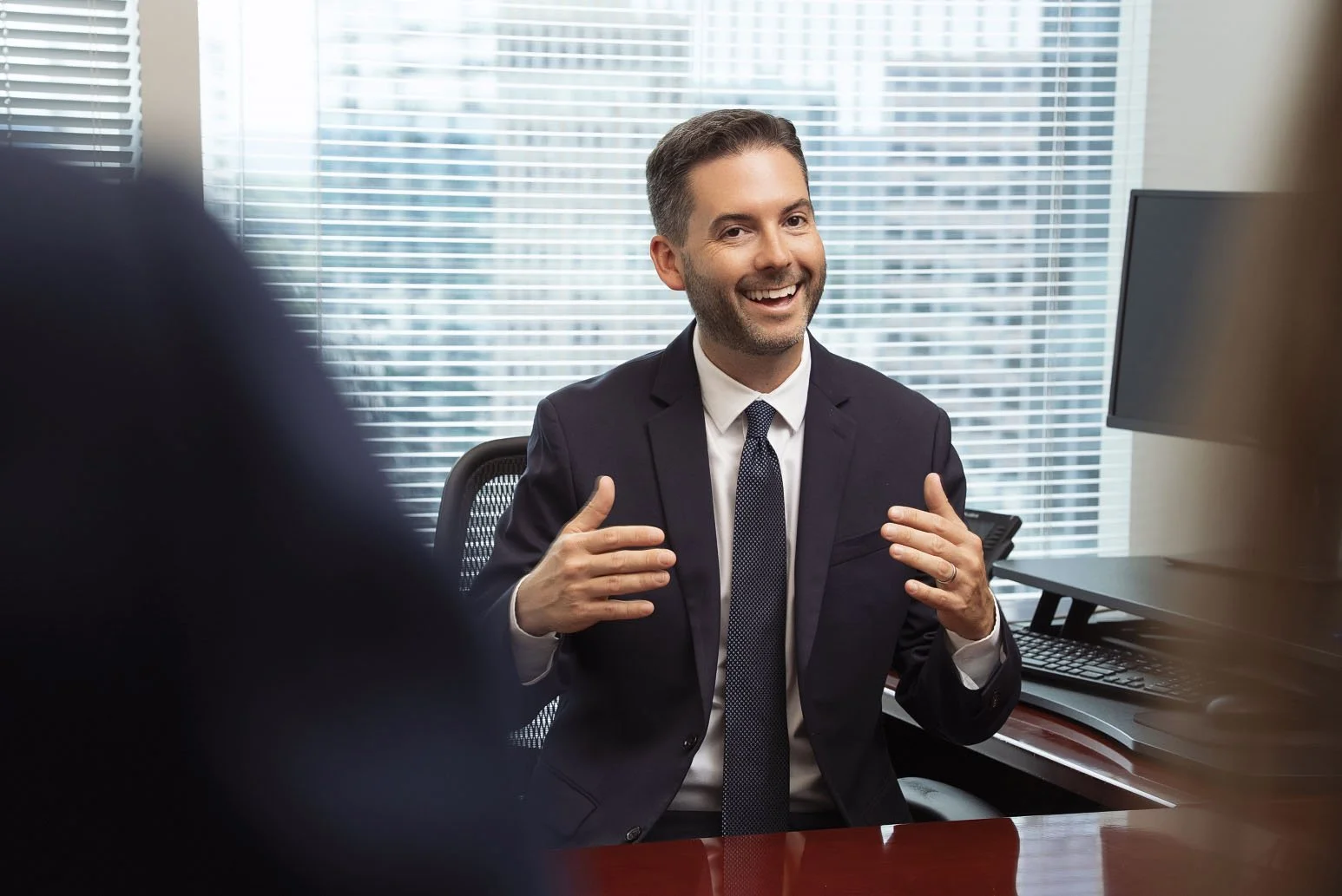 Businessman in a suit smiling and gesturing during a conversation in an office setting.