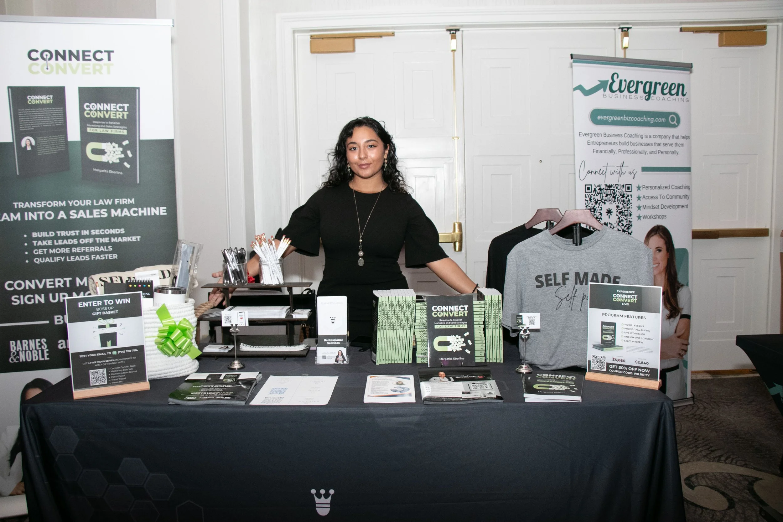 A woman standing behind a black table with promotional materials at an event, with banners for 'Connect & Convert' and 'Evergreen Business Coaching' in the background.