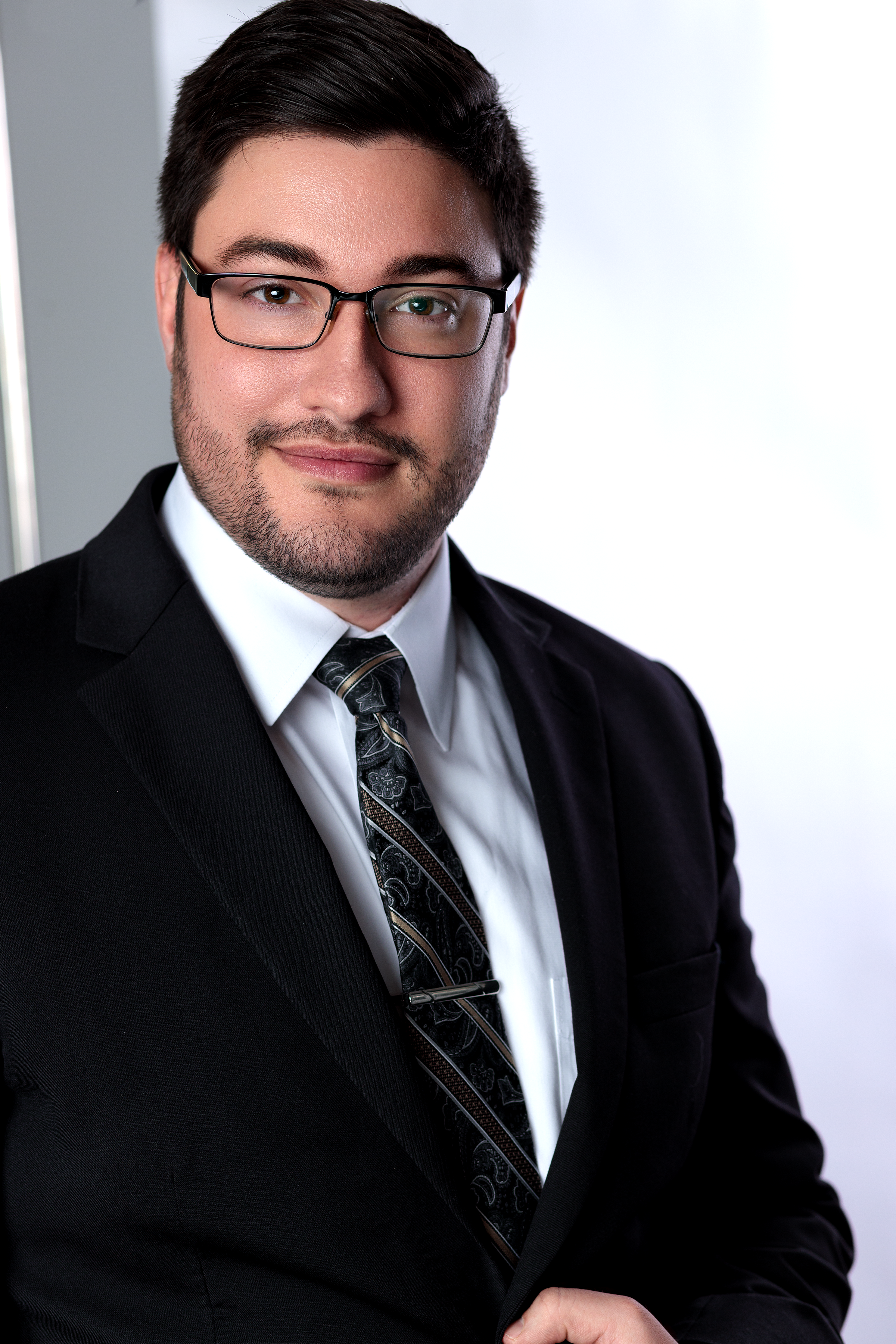 A man in a black suit, white shirt, and patterned tie, wearing glasses, is posing for a professional portrait indoors.
