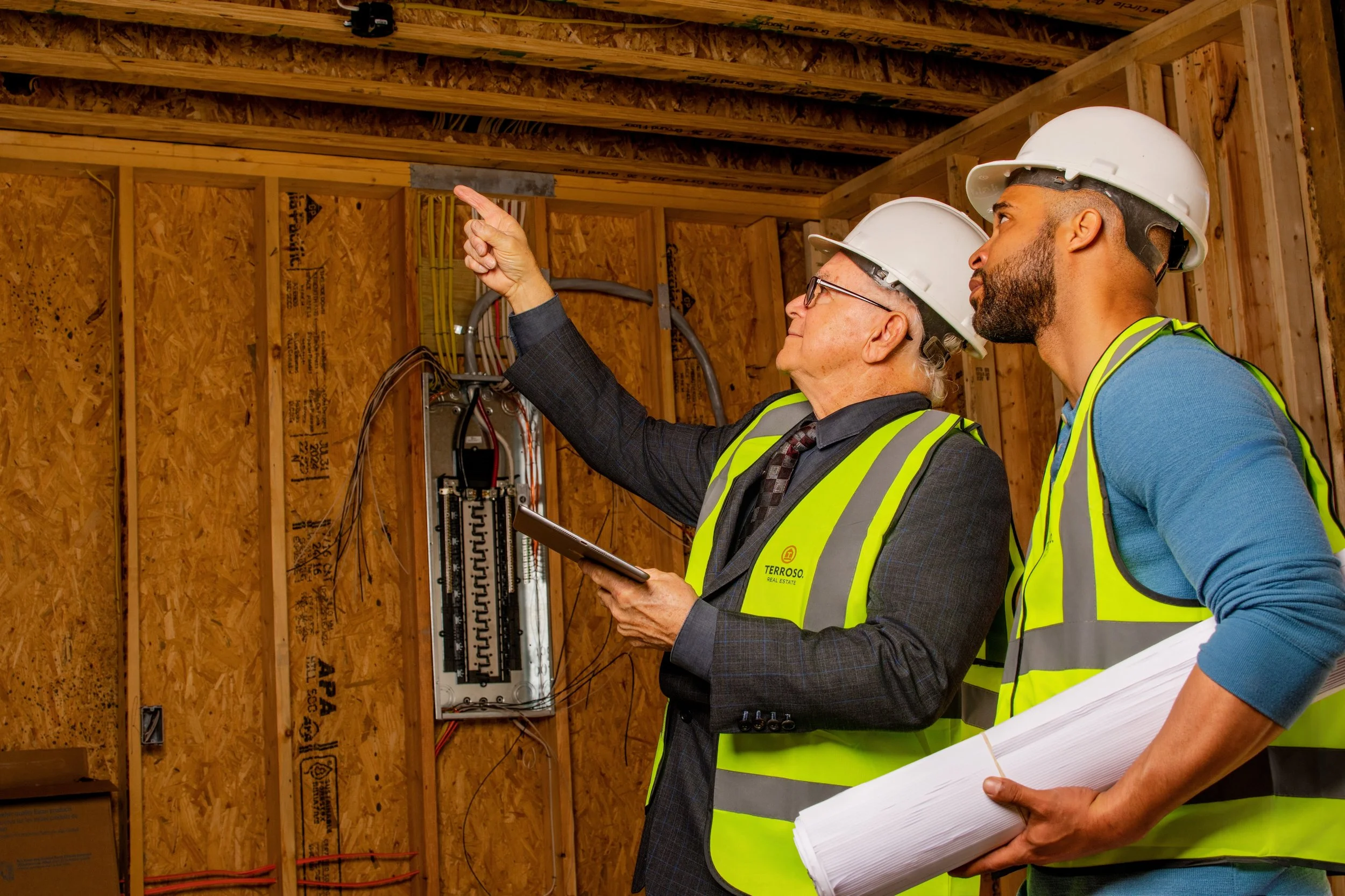 Two construction workers in hard hats and safety vests inspecting electrical work inside a wooden framed structure, with one pointing at the electrical panel and the other holding blueprints.