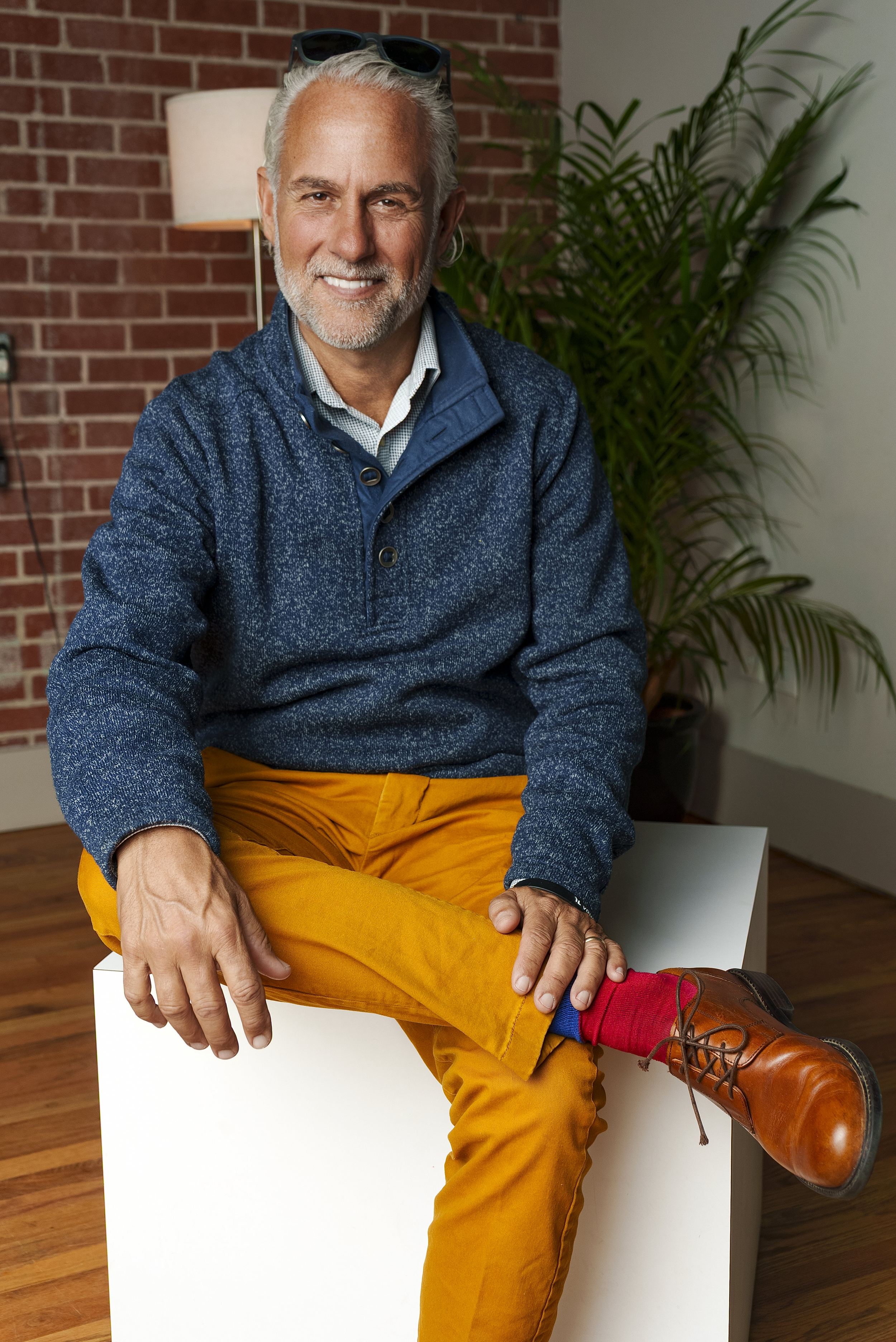 Smiling older man with white hair and beard wearing sunglasses on his head, a blue zip-up sweater, and orange pants, sitting on a white block in a room with brick and white walls and a large potted plant in the background.