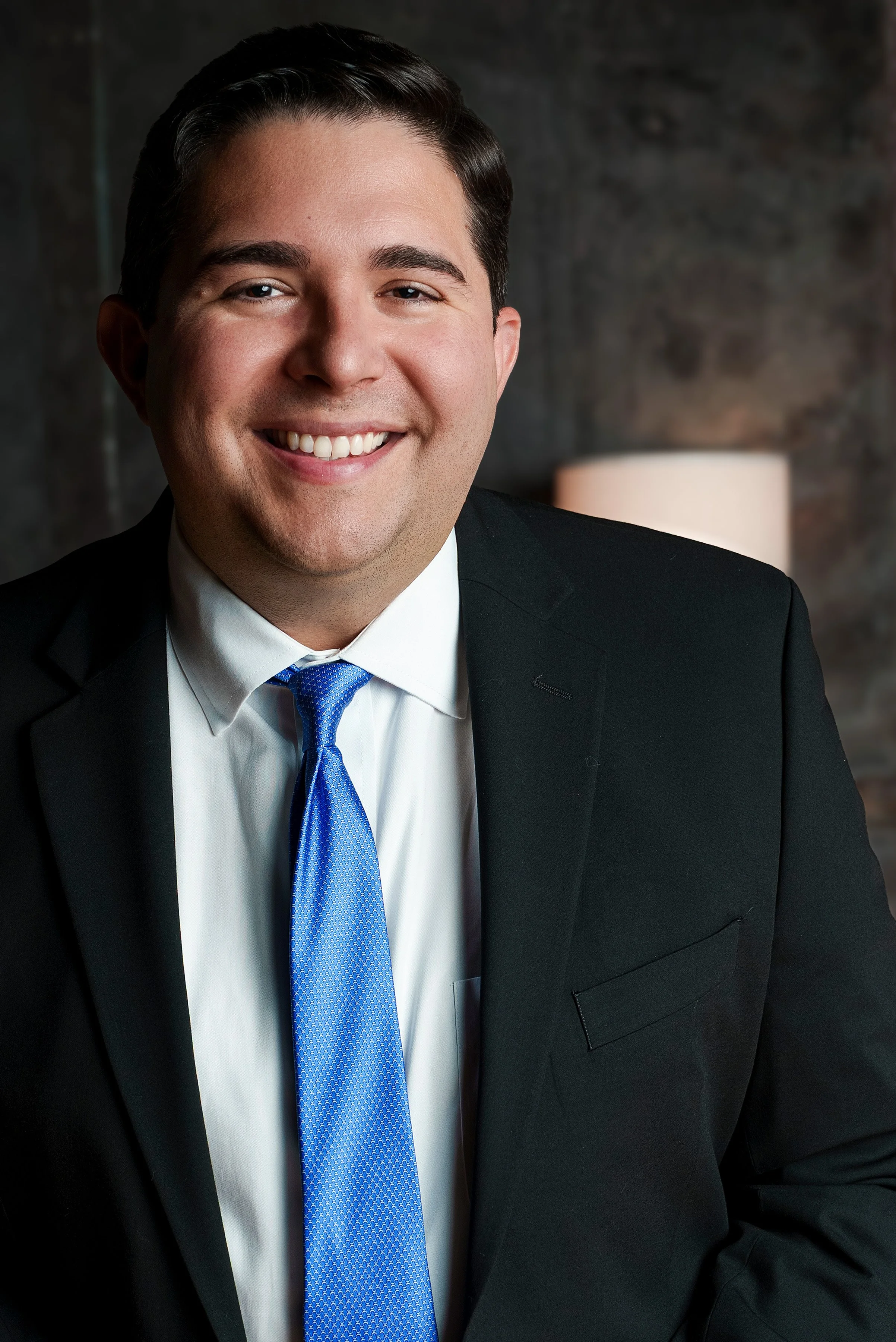 Portrait of a smiling man in a black suit, white shirt, and blue tie, sitting in front of a dark background with a lamp.