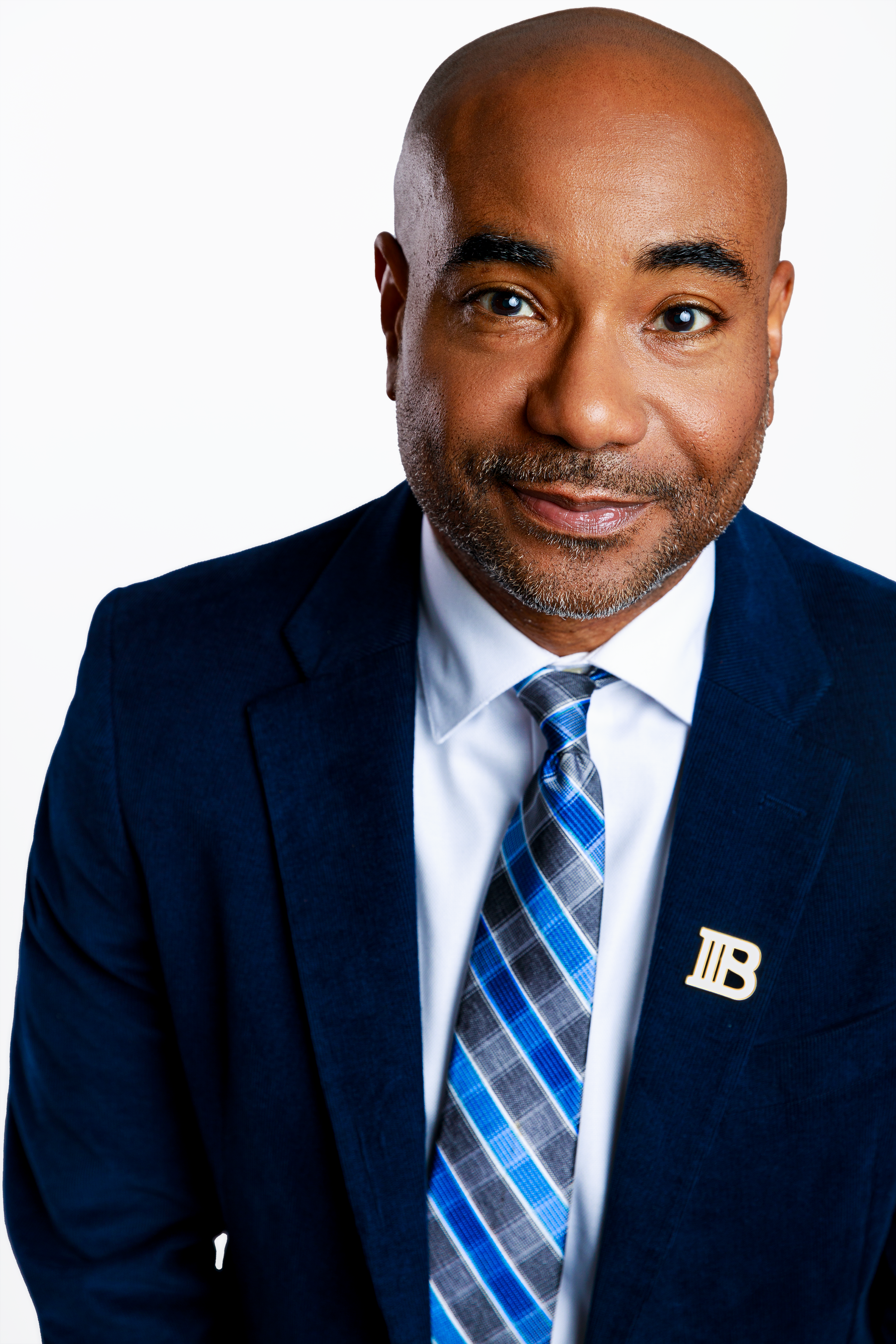 A bald man with a beard wearing a dark blue suit, white shirt, and a blue plaid tie, smiling at the camera against a white background.