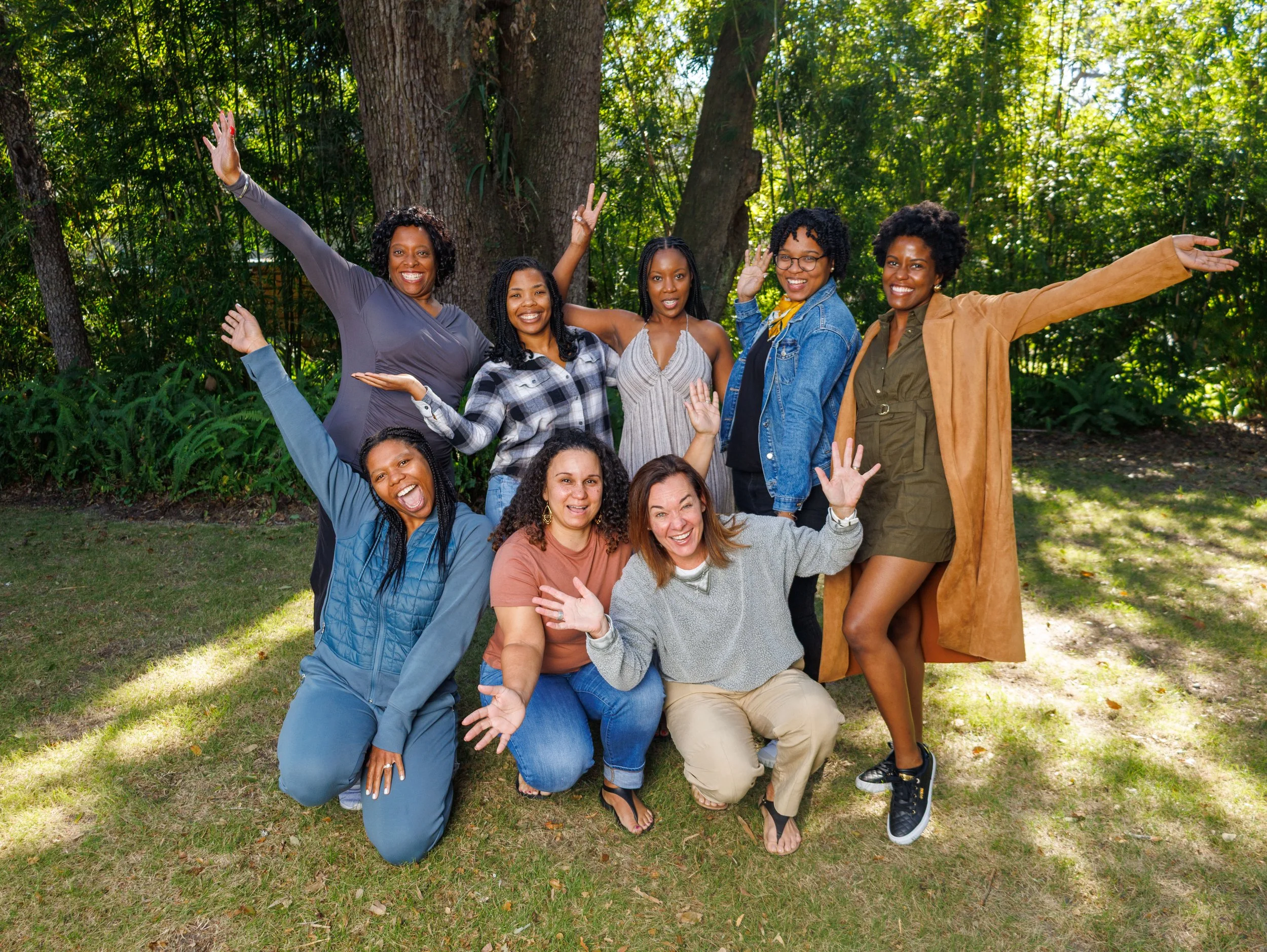 A group of ten diverse women happily posing outdoors in a park, standing and kneeling on a grassy area with trees in the background.