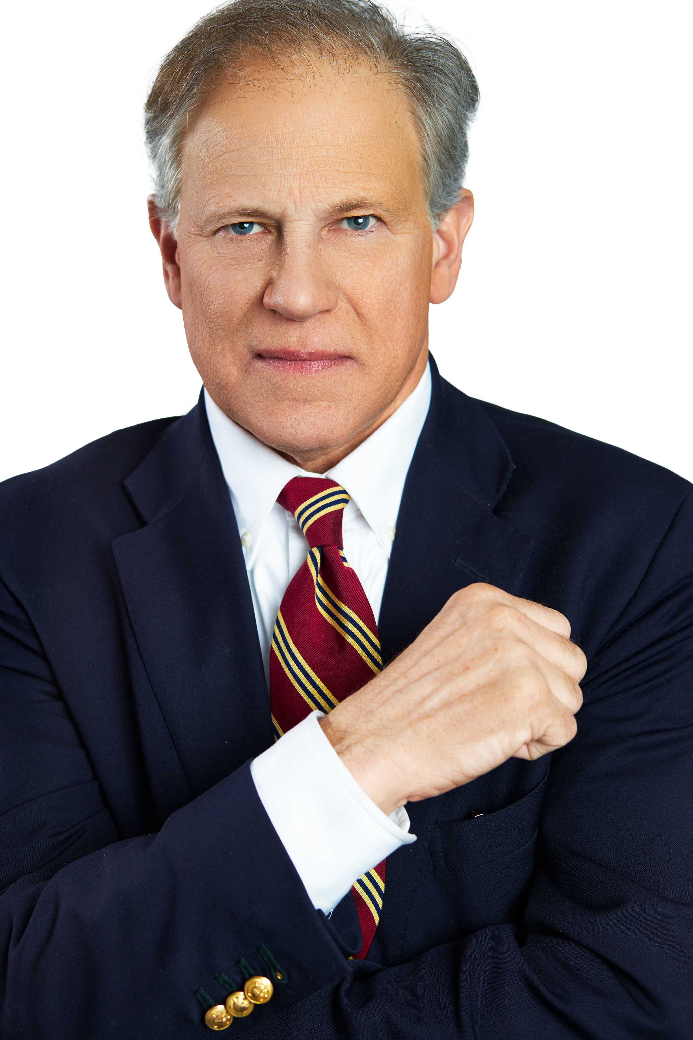 A confident middle-aged man with gray hair and blue eyes, dressed in a dark navy suit, white shirt, and striped red and yellow tie, posing with his arms crossed against a white background.