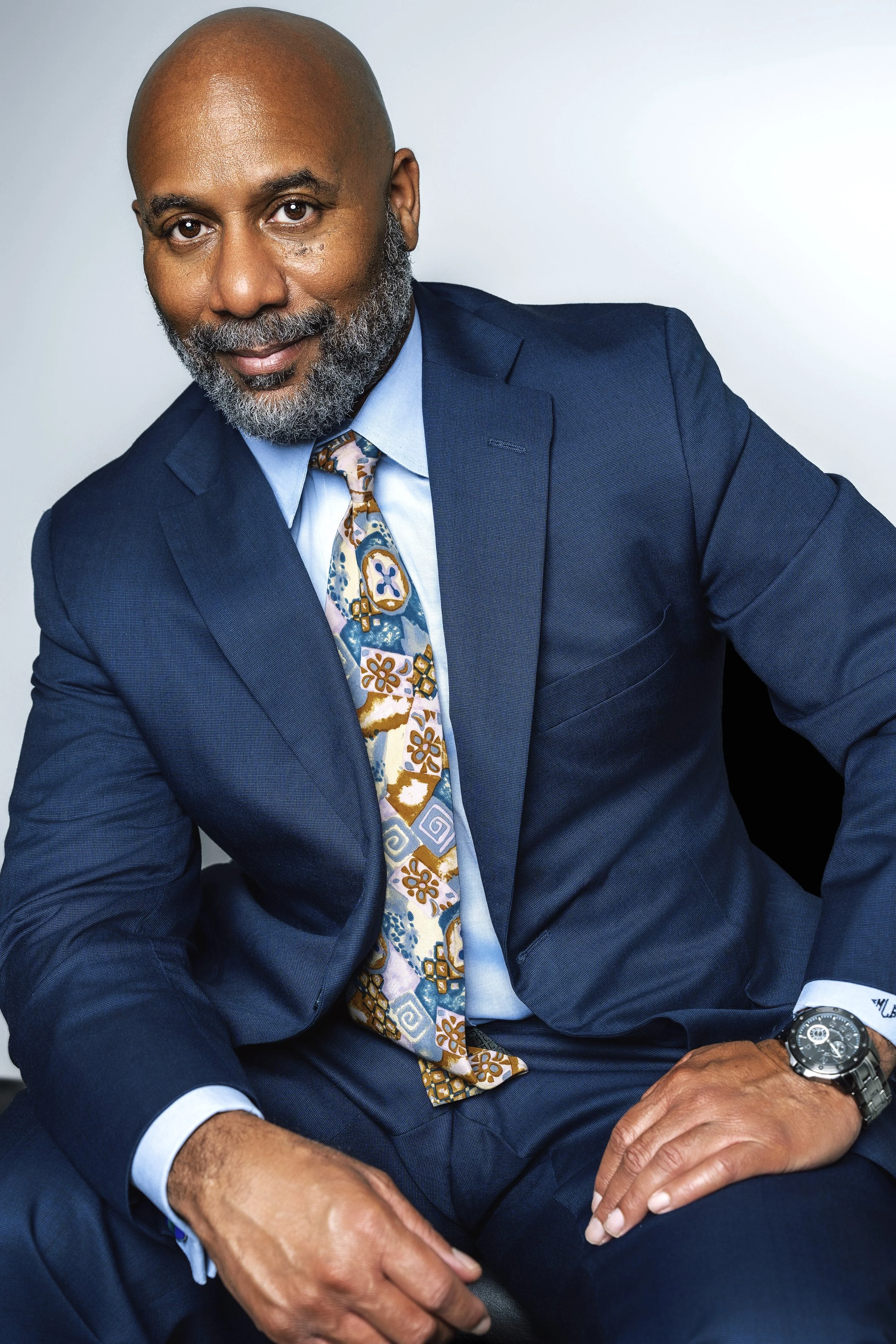 A middle-aged African American man in a blue suit, light blue shirt, and patterned tie, sitting against a light gray background. He is smiling and looking at the camera.