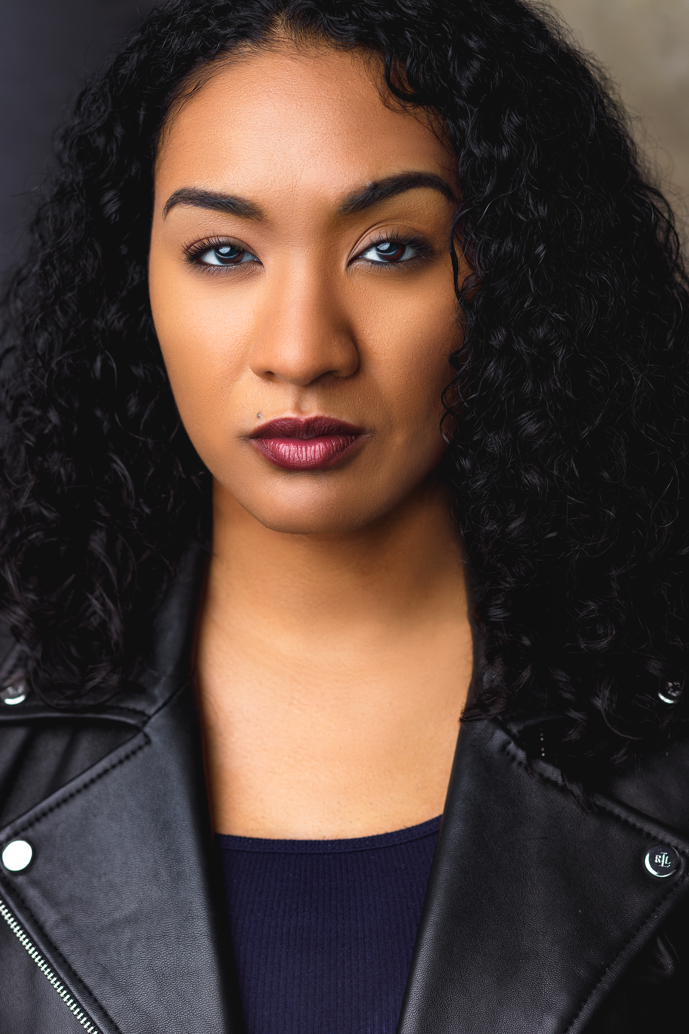 A woman with dark, curly hair and blue eyes wearing a black leather jacket and dark lipstick.