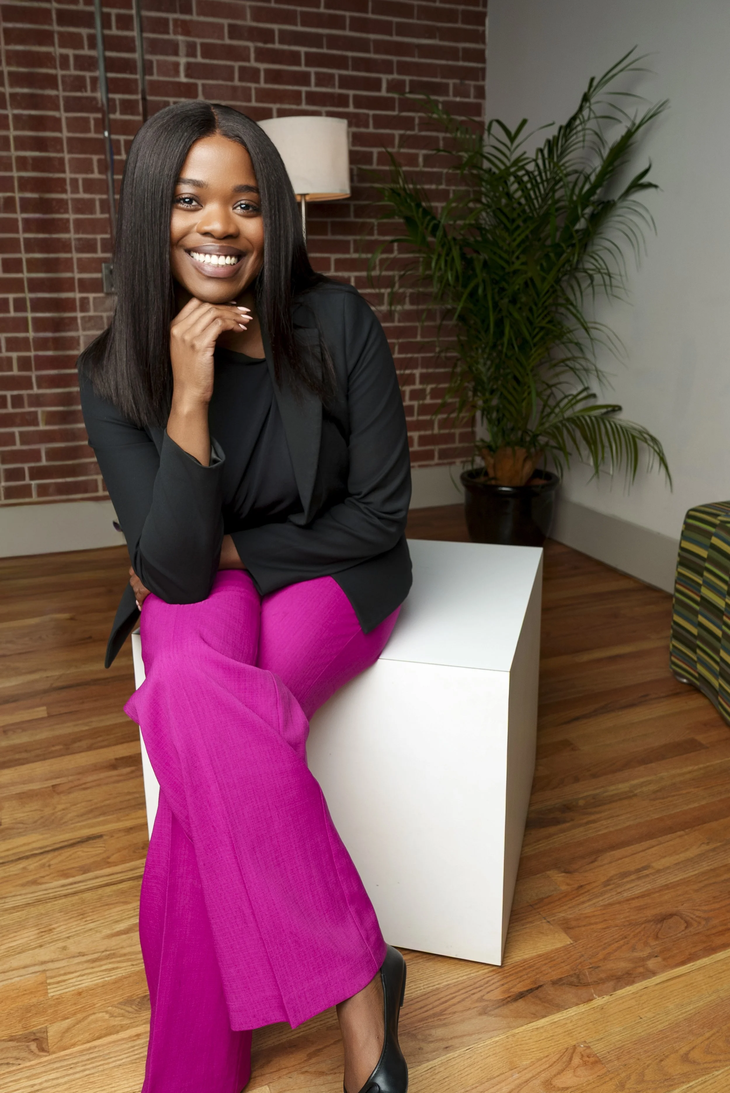 A woman with dark hair, smiling, wearing a black blazer and bright pink pants, sitting on a white block in a room with a brick wall, a potted plant, and a striped armchair in the background.