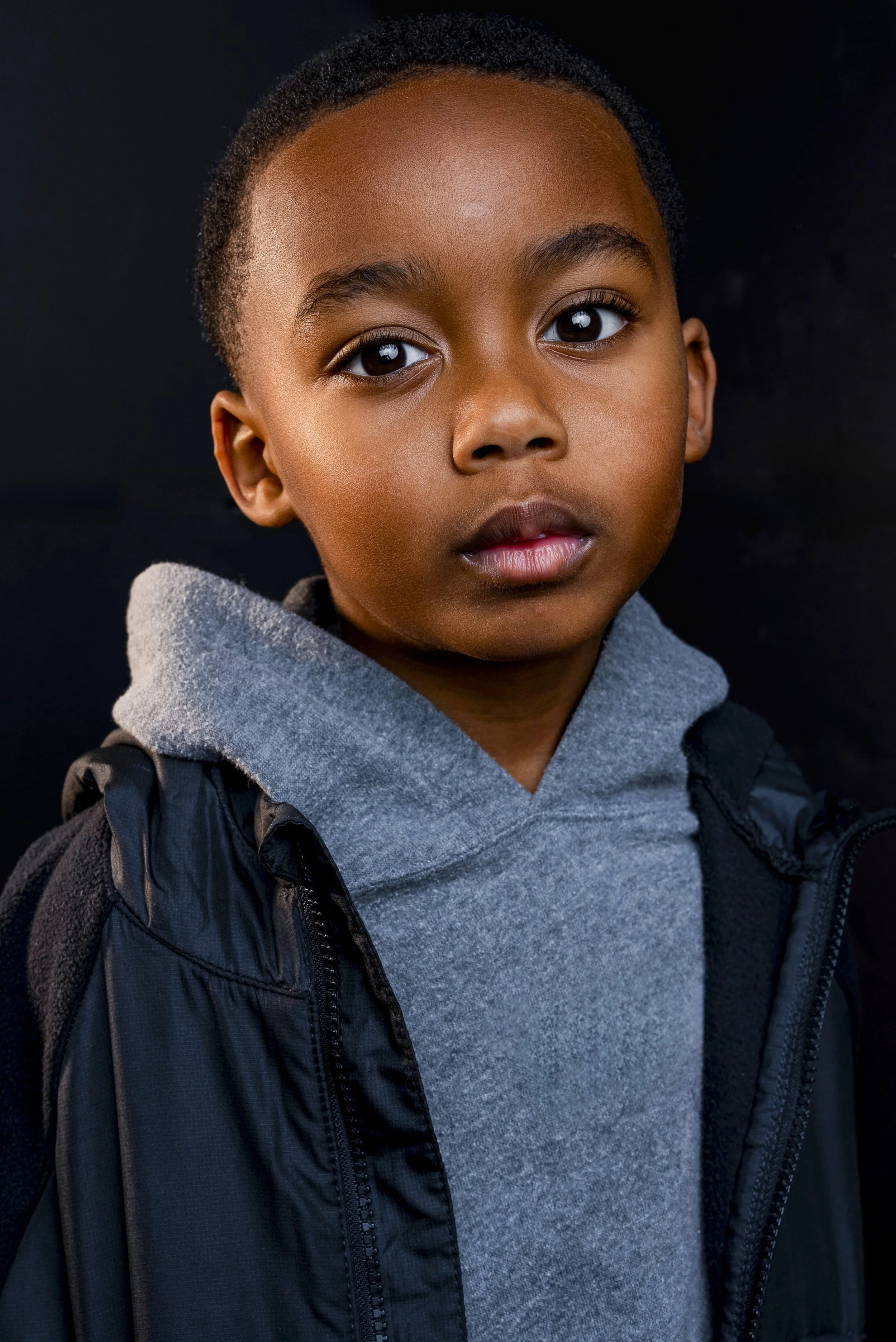 Close-up of a young boy with short curly hair, wearing a gray hoodie and black jacket, against a dark background.