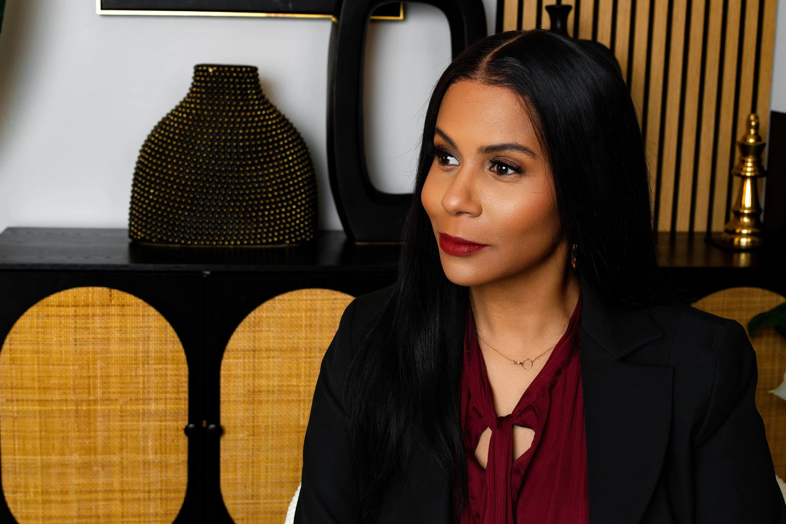 A woman with long black hair, wearing a black blazer and a red blouse, sitting indoors with a modern, decorative background including a black and gold vase, a black sculpture, and wooden paneling.