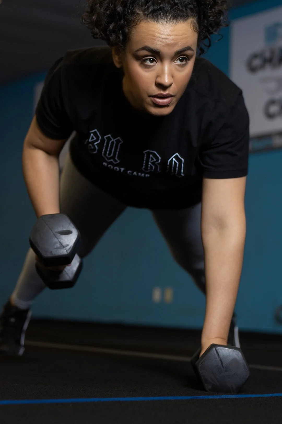 A woman with curly hair doing a plank exercise with dumbbells in a gym.