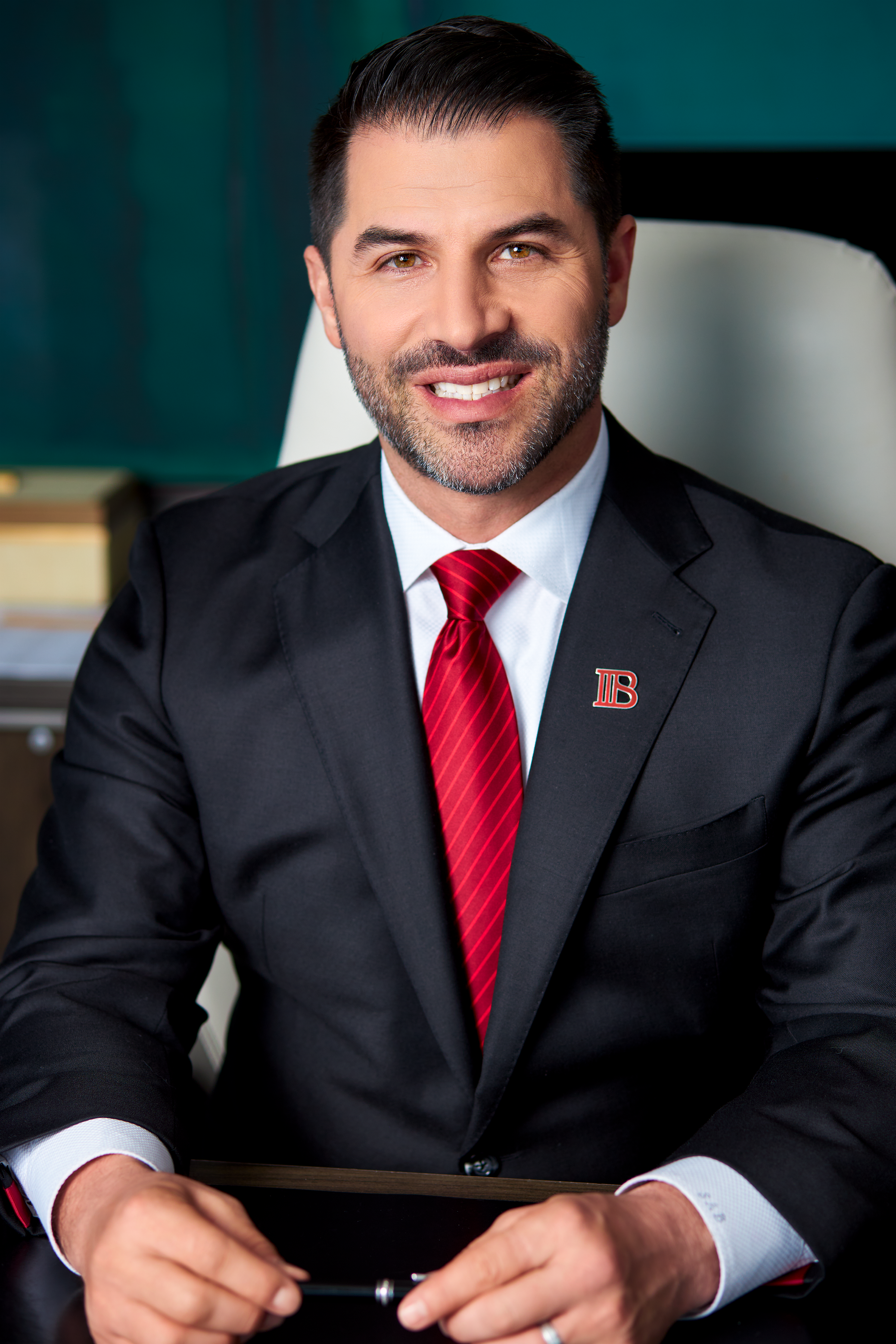 A man in a suit and red tie sitting at a desk in an office, smiling at the camera.