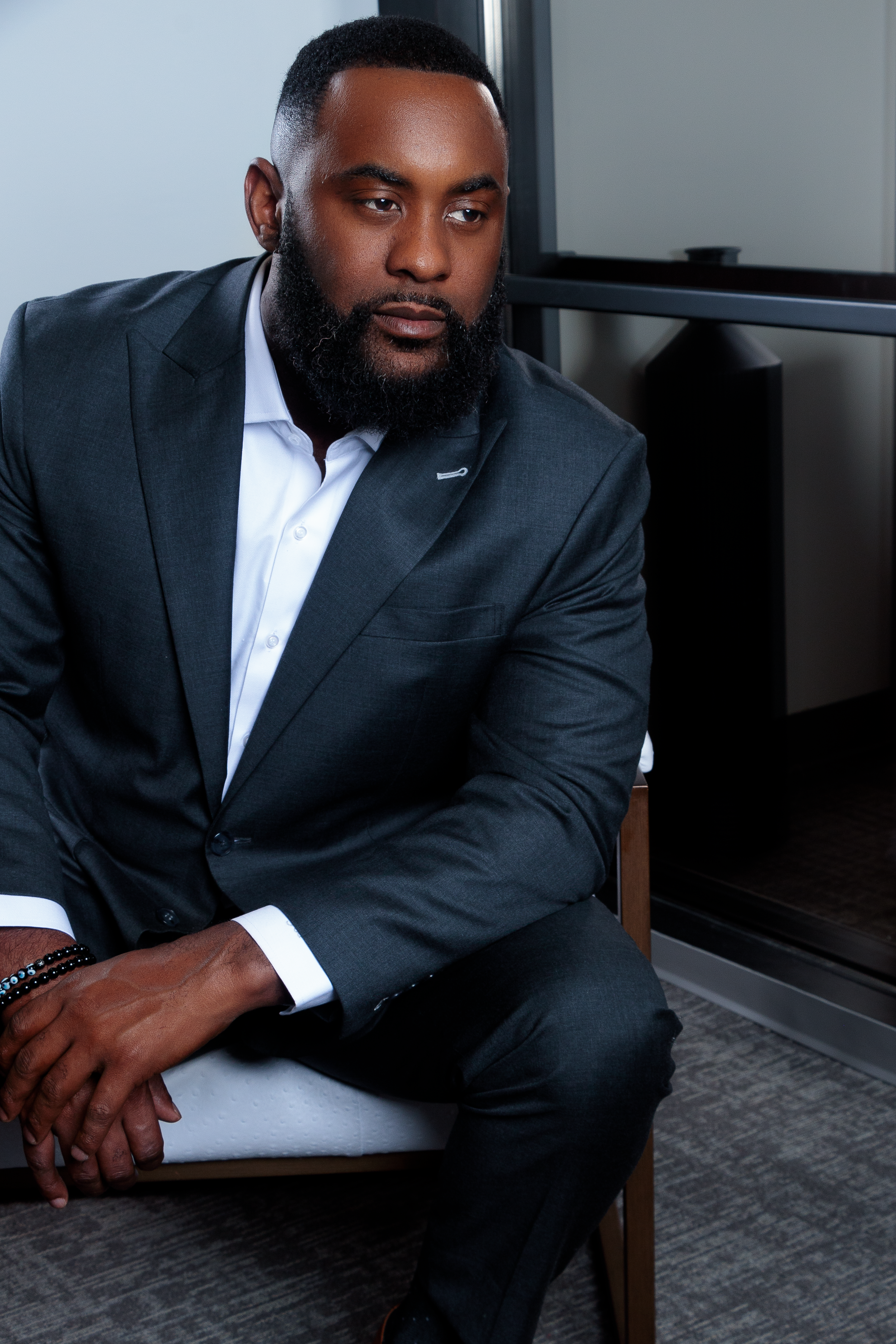 A man in a dark gray suit and white shirt sitting on a chair near a window in an office.