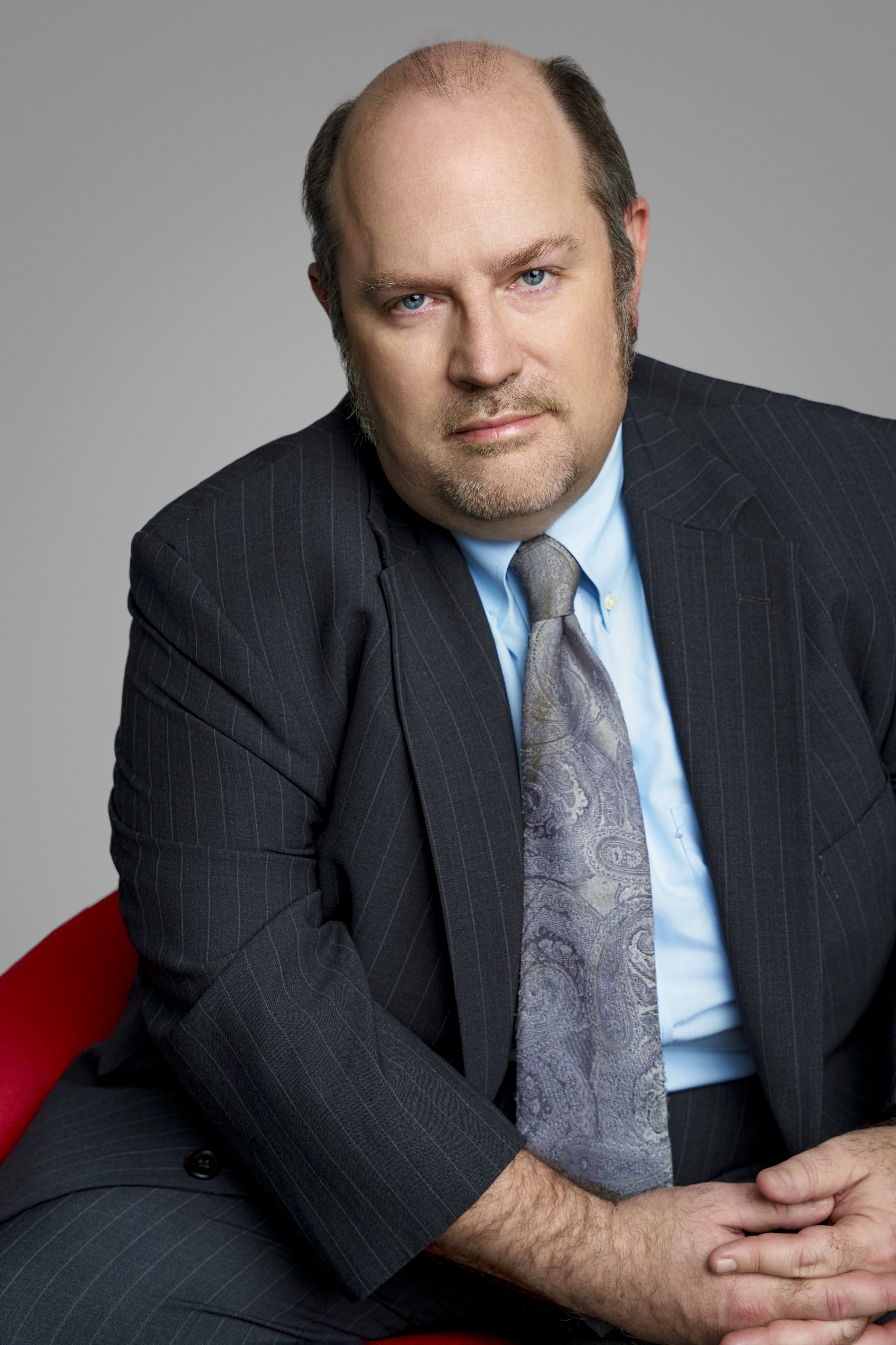 A middle-aged man with light skin, blue eyes, and a bald head with hair on the sides. He is wearing a dark pinstripe suit, a light blue dress shirt, and a paisley gray tie. He is seated on a red chair or sofa, with his hands clasped together. The bac