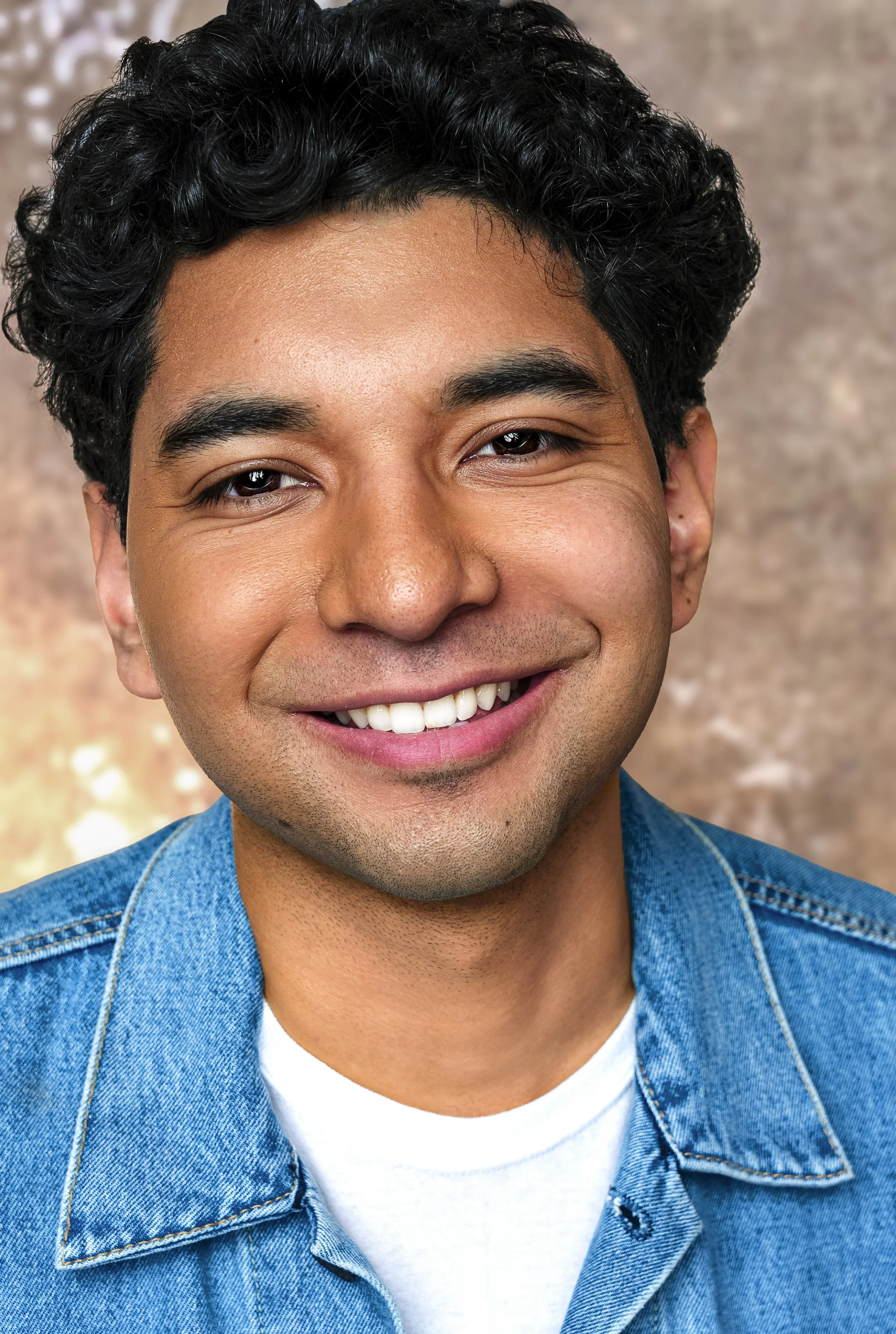 Close-up portrait of a smiling young man with dark curly hair, wearing a denim jacket over a white shirt, against a softly blurred brown background.