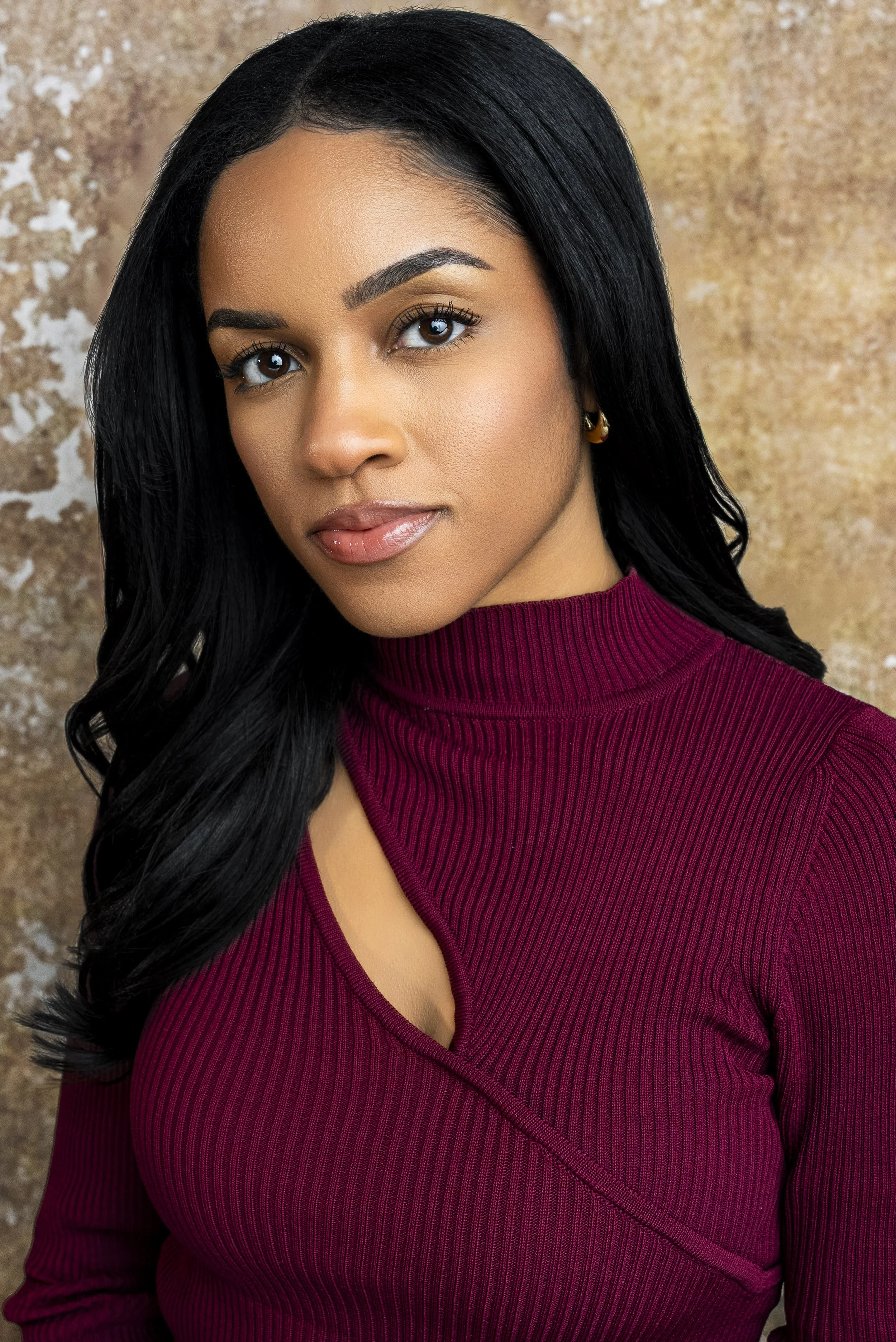 A woman with long black hair, wearing a burgundy ribbed turtleneck sweater with a cutout detail, standing against a textured beige background.