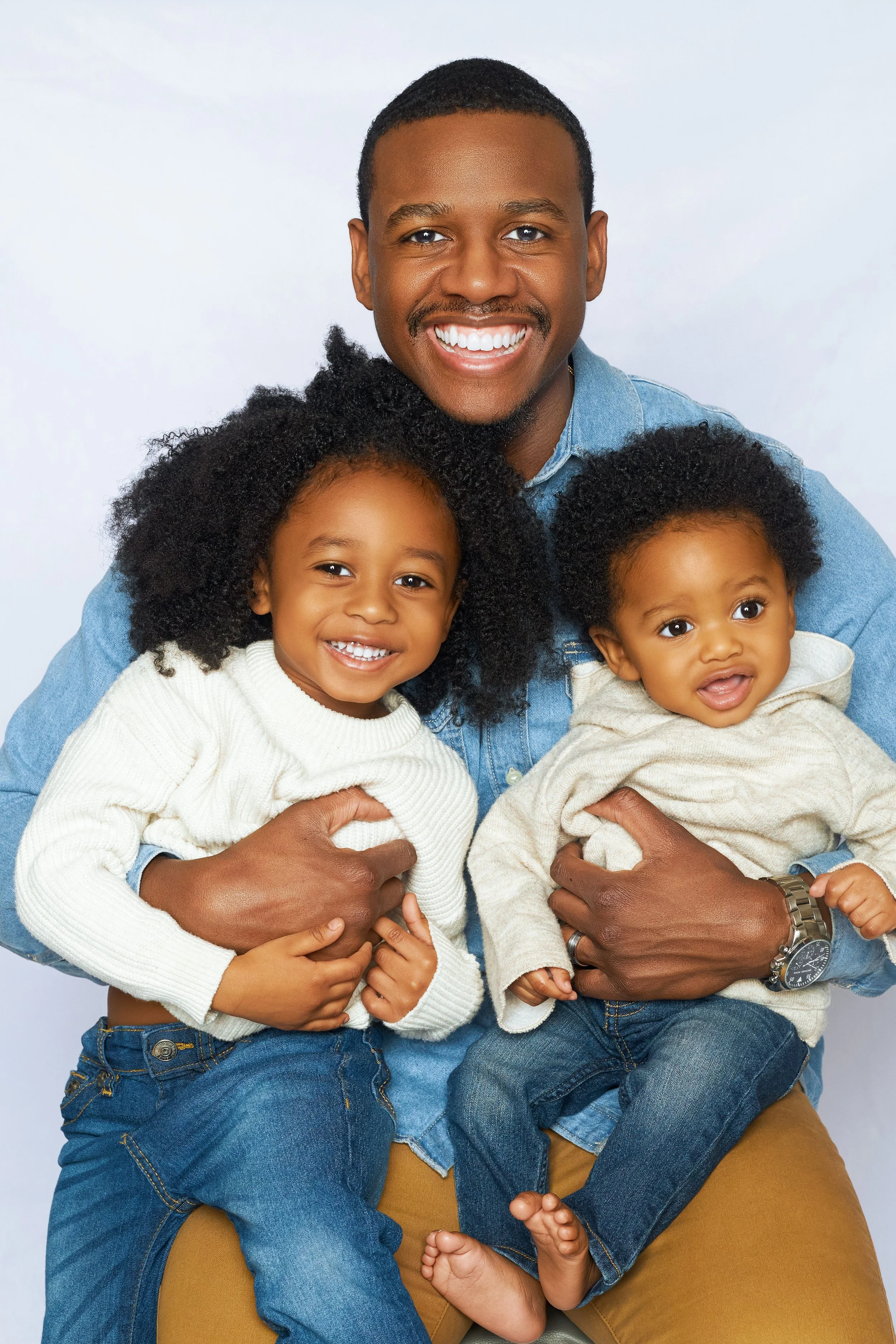 A smiling man holding two young children, one in each arm, against a white background.