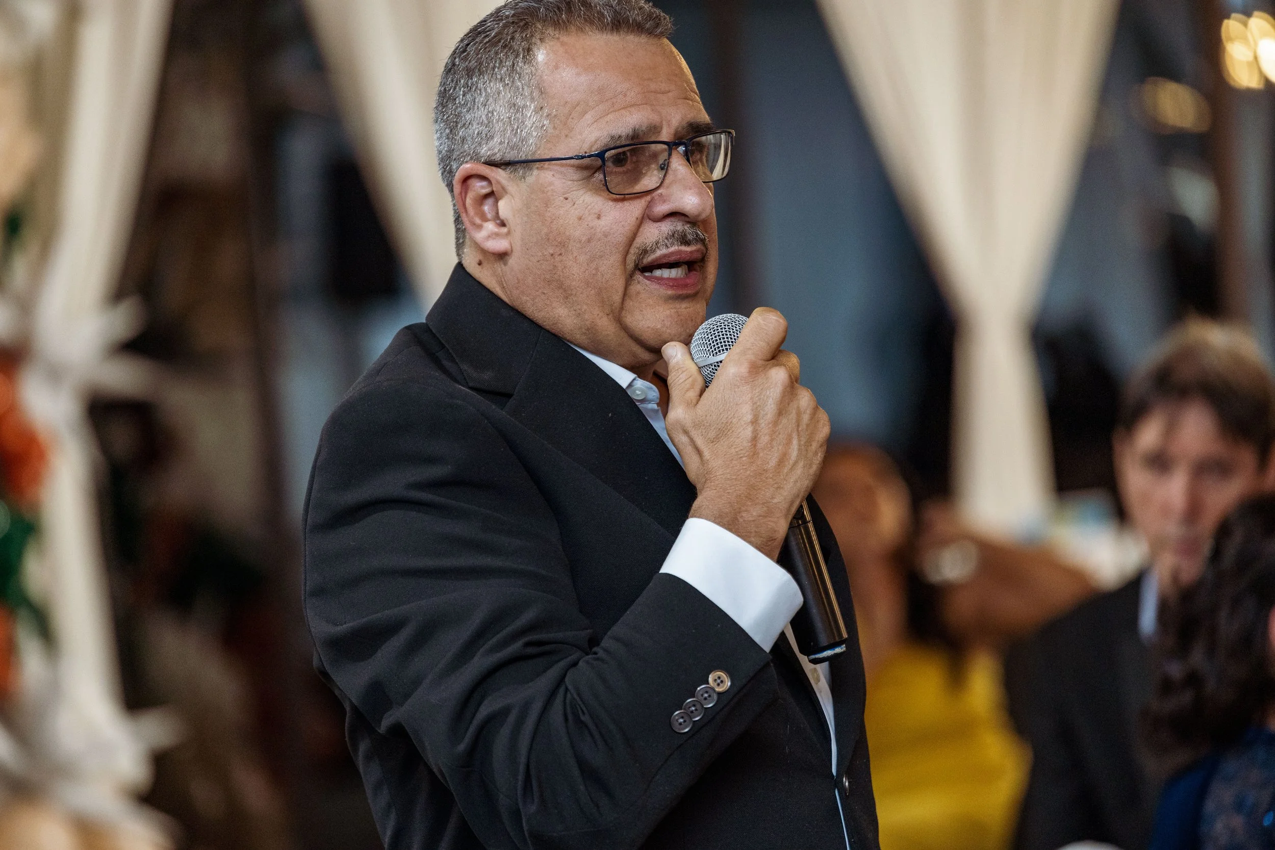 Man wearing glasses, black suit, and white shirt speaking into a microphone at an indoor event.