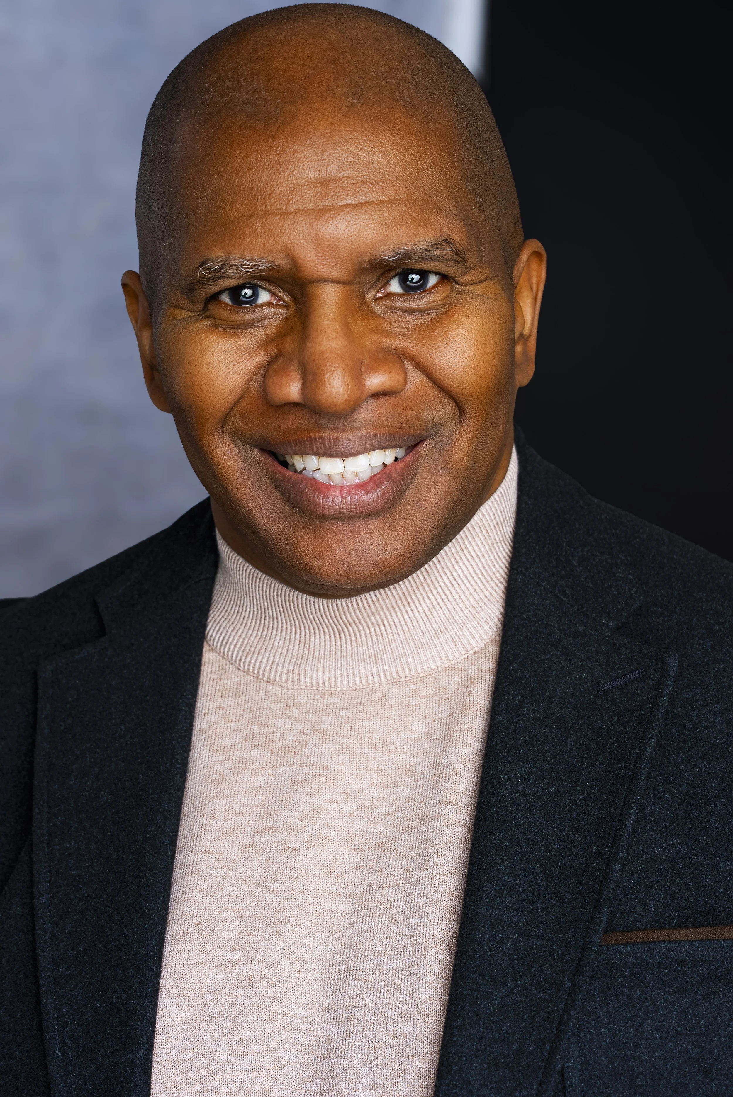 Close-up portrait of a smiling Black man with blue eyes, wearing a beige turtleneck and a dark blazer.