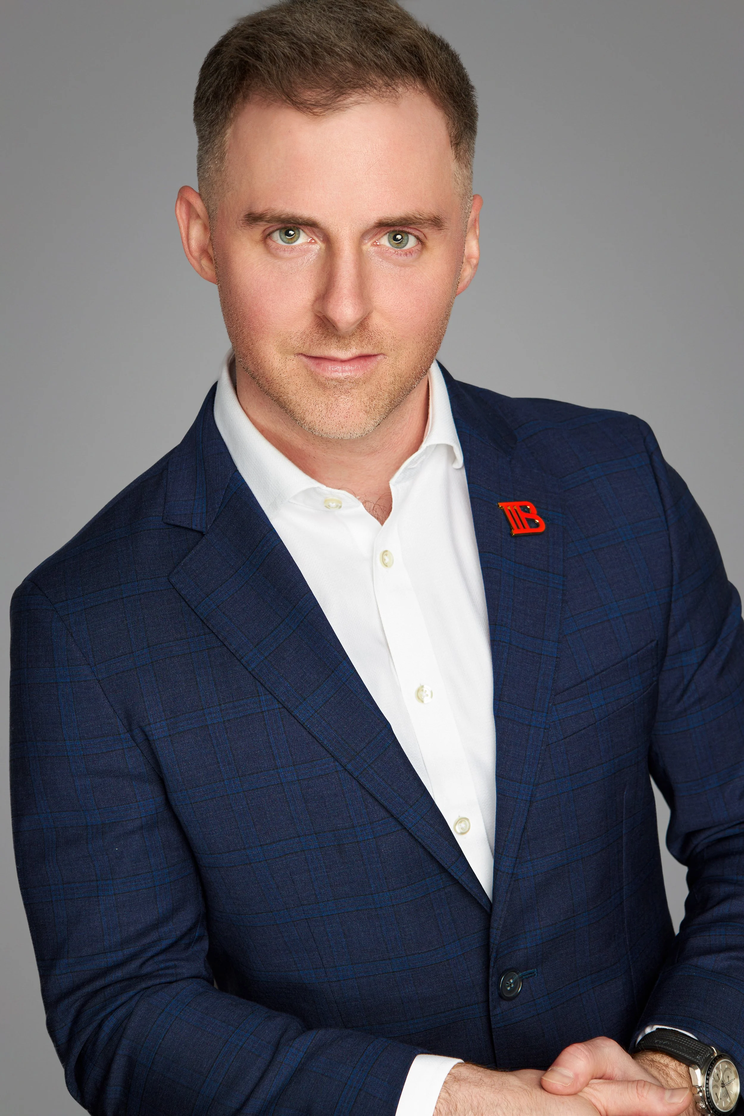 A man with short brown hair and green eyes wearing a dark blue plaid suit with a white shirt, posing against a gray background.