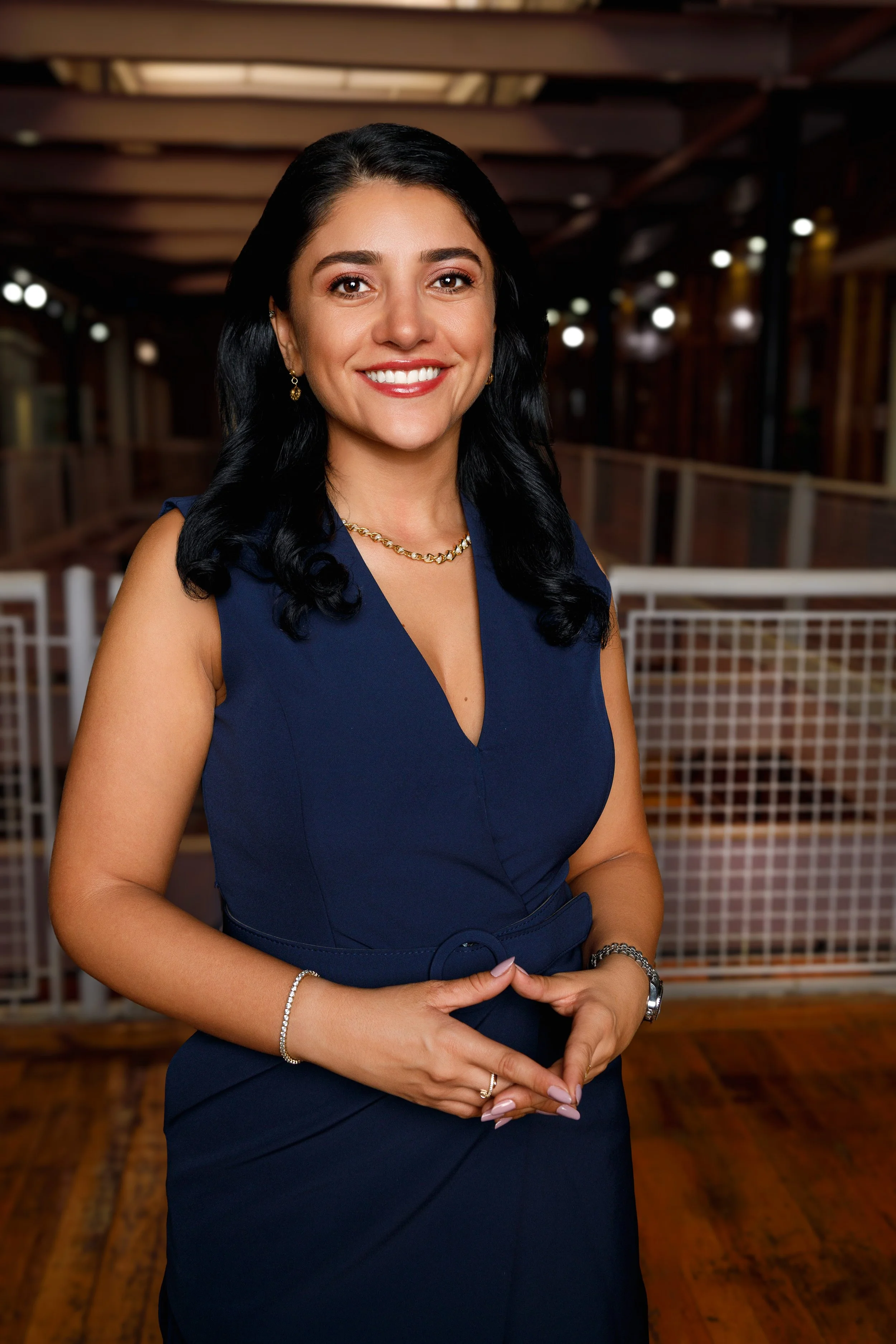A woman with black hair, wearing a navy blue sleeveless dress, smiling and standing indoors with wooden floors and fencing in the background.