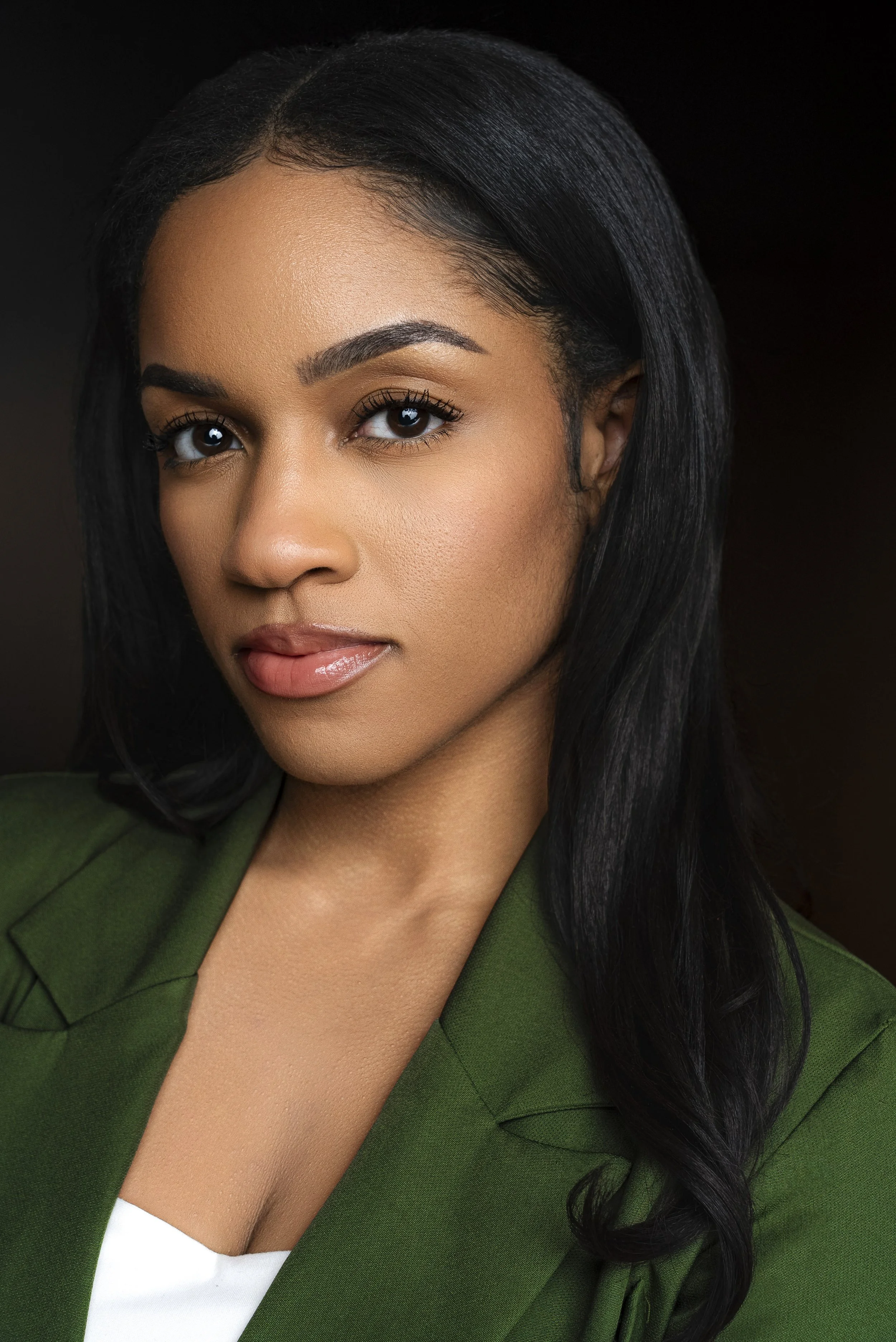 A close-up portrait of a woman with black hair, brown eyes, and a light makeup look, wearing a green blazer over a white top, against a dark background.