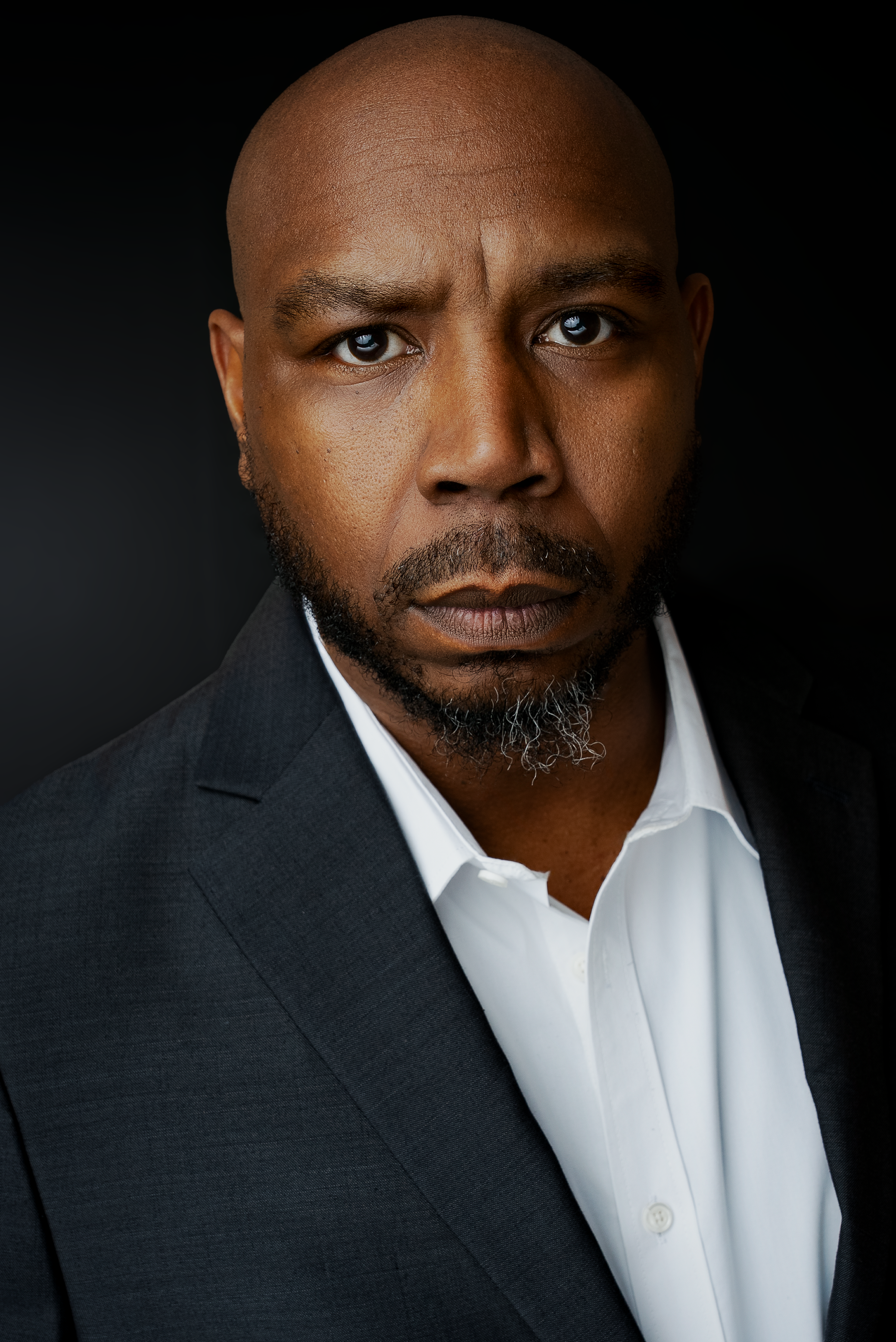 Close-up portrait of a serious African American man with a beard, wearing a black suit and white shirt, against a dark background.