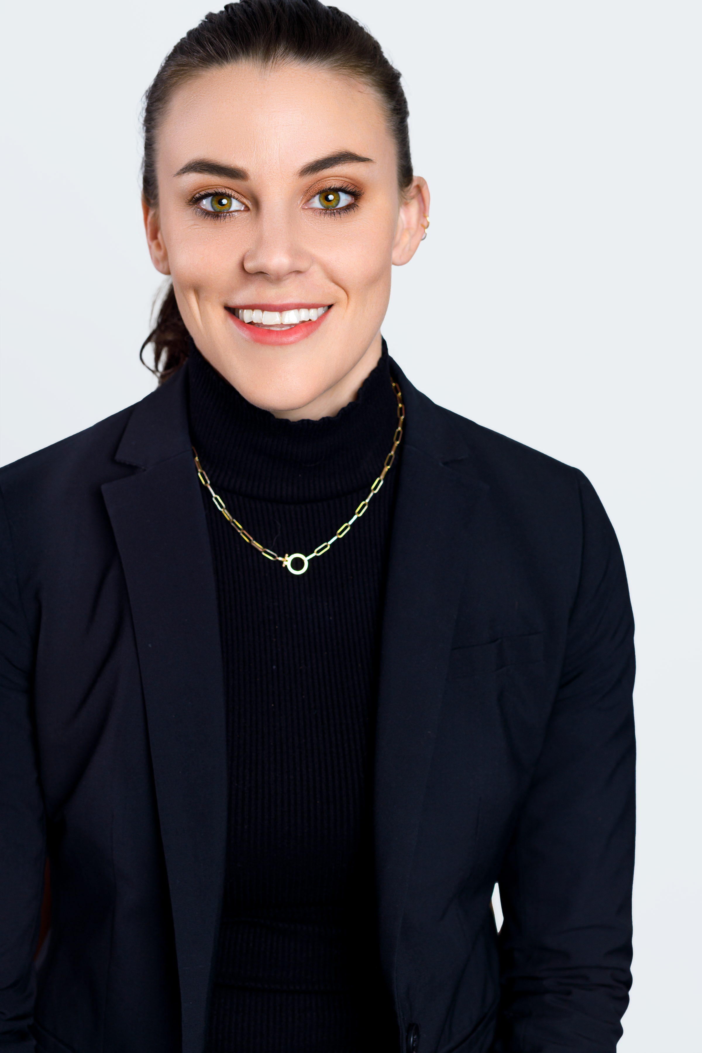 A woman with brown hair pulled back, wearing a black turtleneck and blazer, smiling at the camera, with a gold chain necklace, against a light gray background.