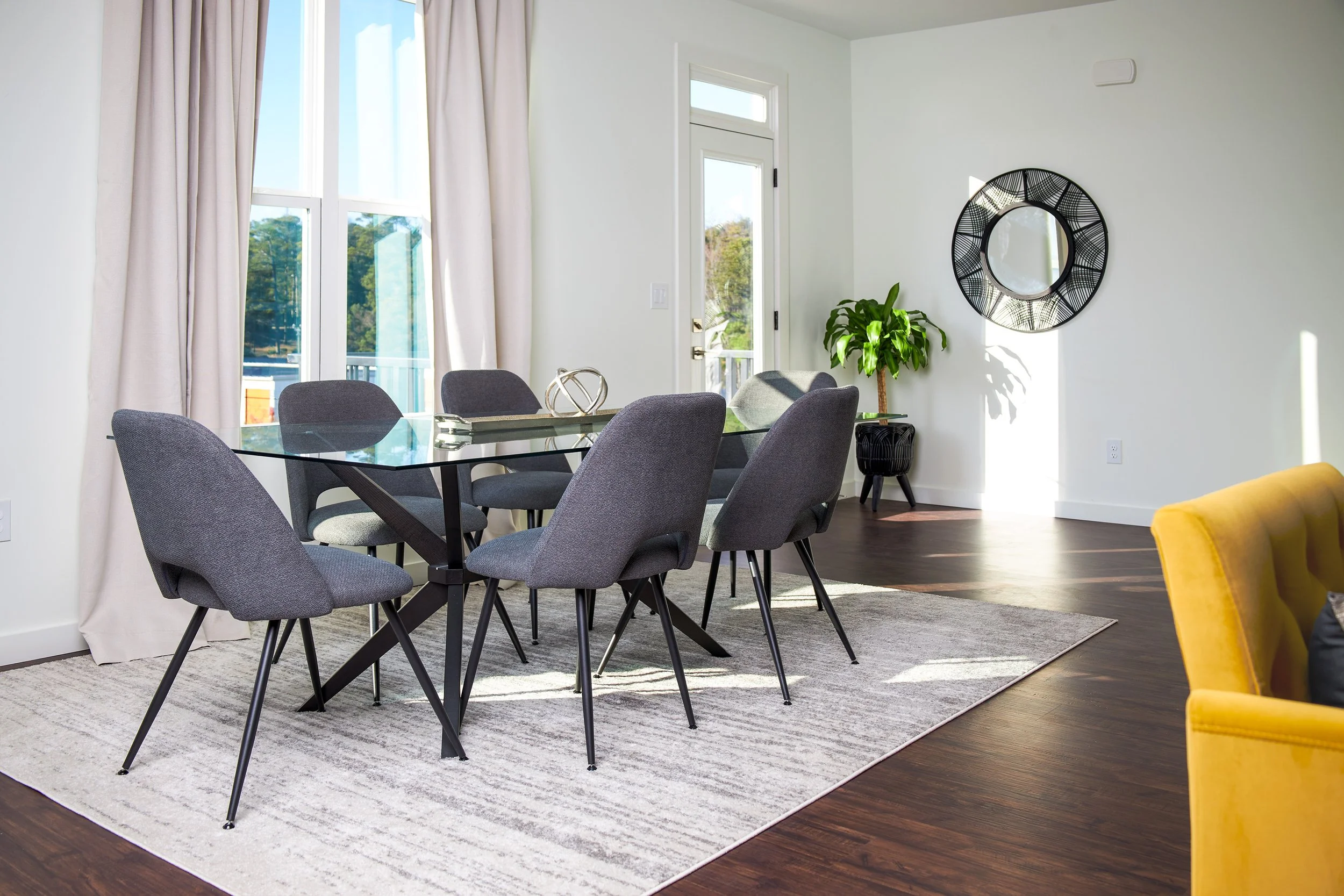 A dining room with a glass-top table surrounded by six gray upholstered chairs, a white and gray patterned rug underneath, a large window with beige curtains, a door leading outside, a round black decorative mirror and a green potted plant on a stand