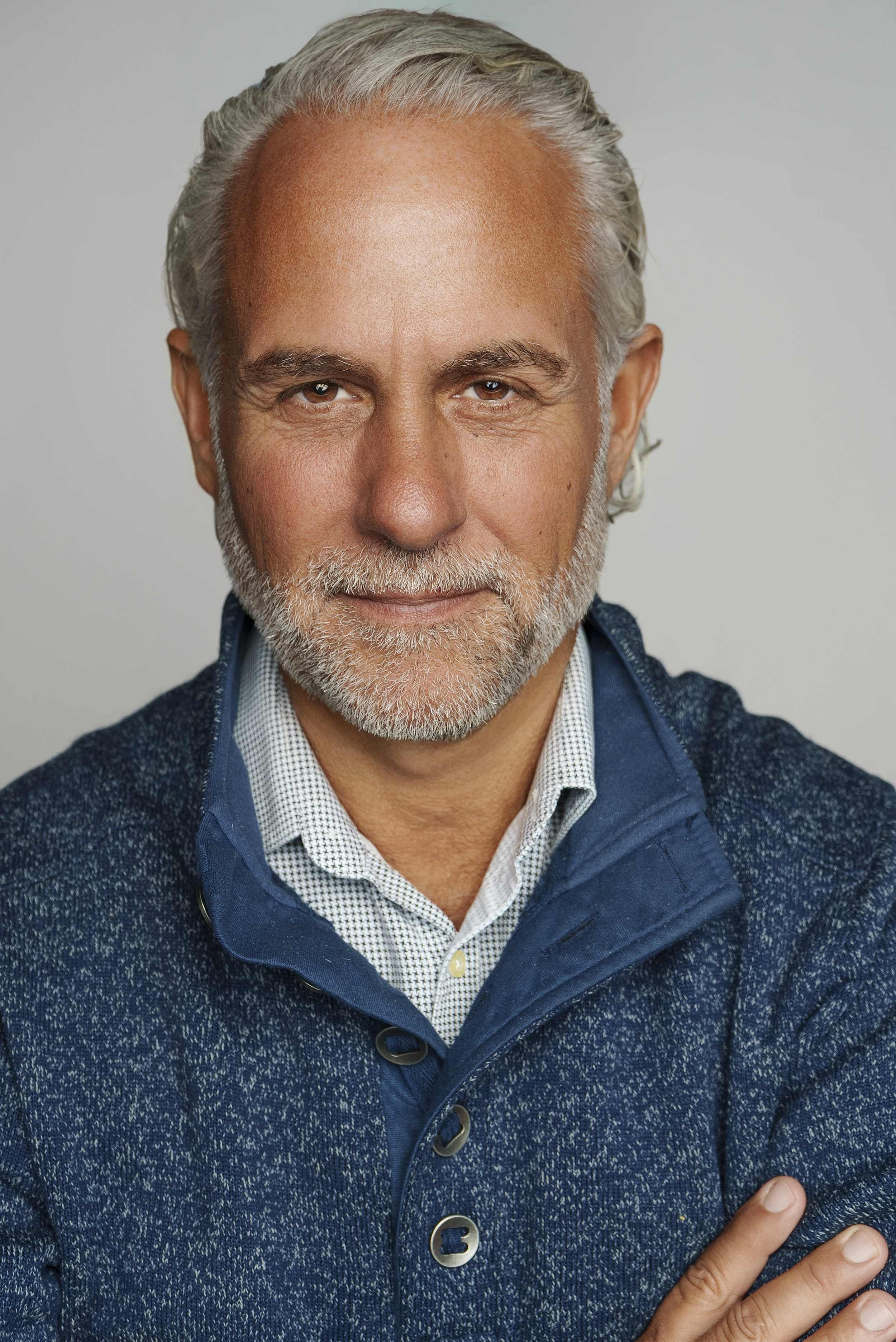 A middle-aged man with gray hair and a beard, wearing a blue zip-up sweater and a collared shirt, looking directly at the camera with a slight smile.