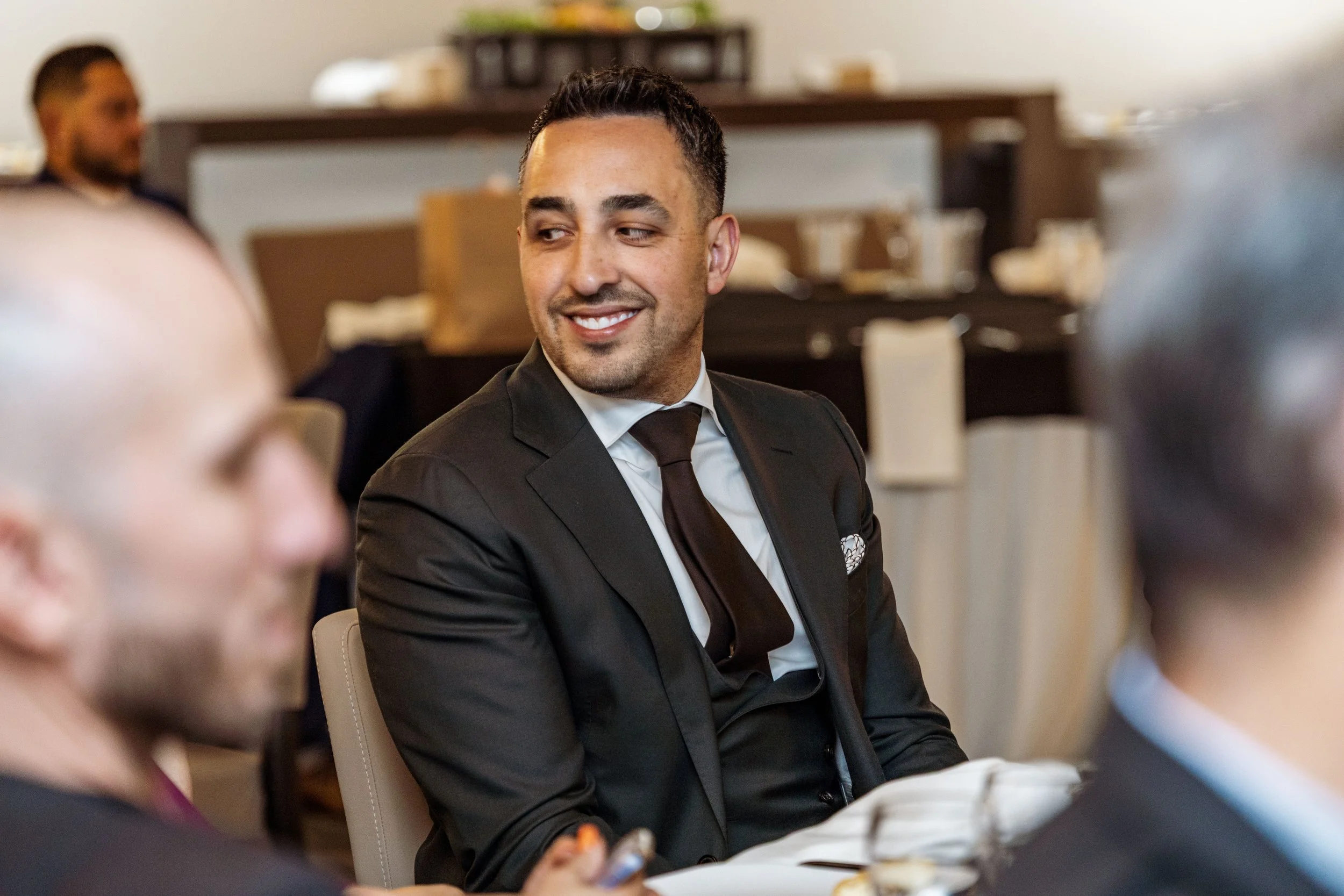 A man in a black suit, white shirt, and black tie, smiling and looking to his right at a formal event or business meeting.